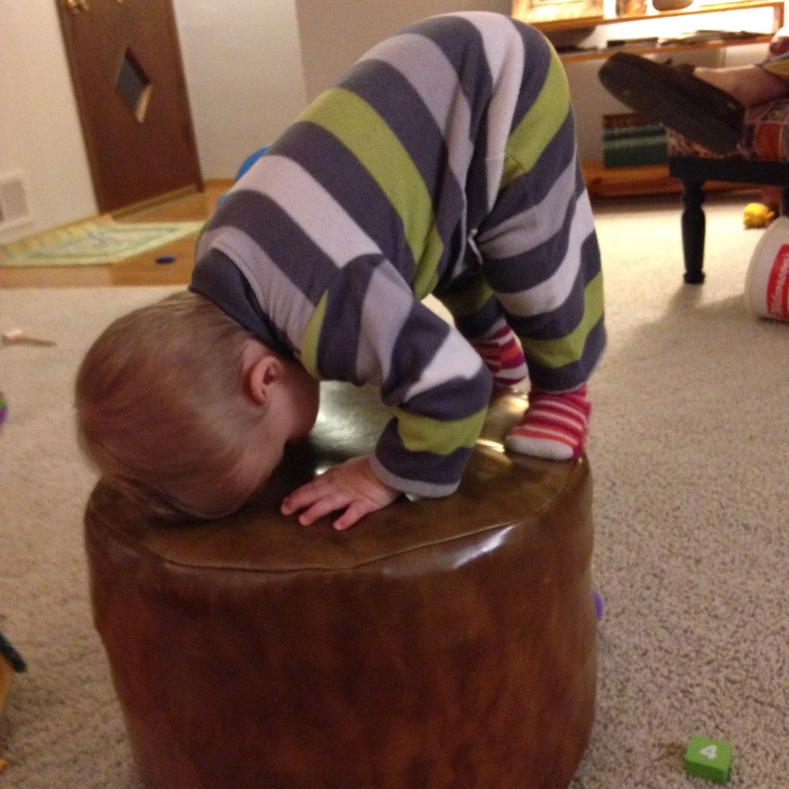 A young child in striped pajamas and socks kneeling on a leather ottoman with their head down and hand on the surface, in a living room.