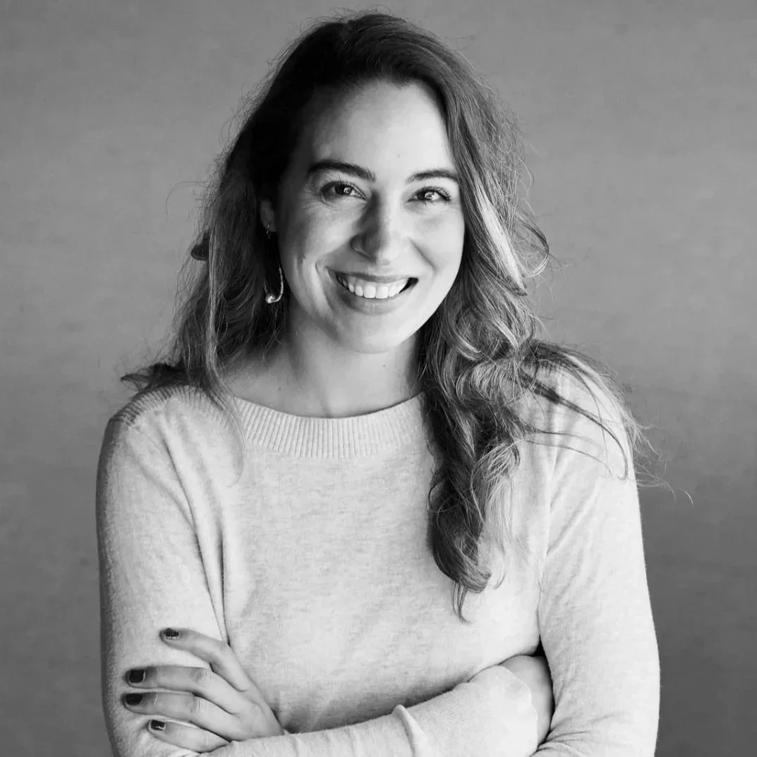 Black and white portrait of a woman with long wavy hair, smiling with arms crossed, wearing a light-colored sweater and earrings, against a plain background.