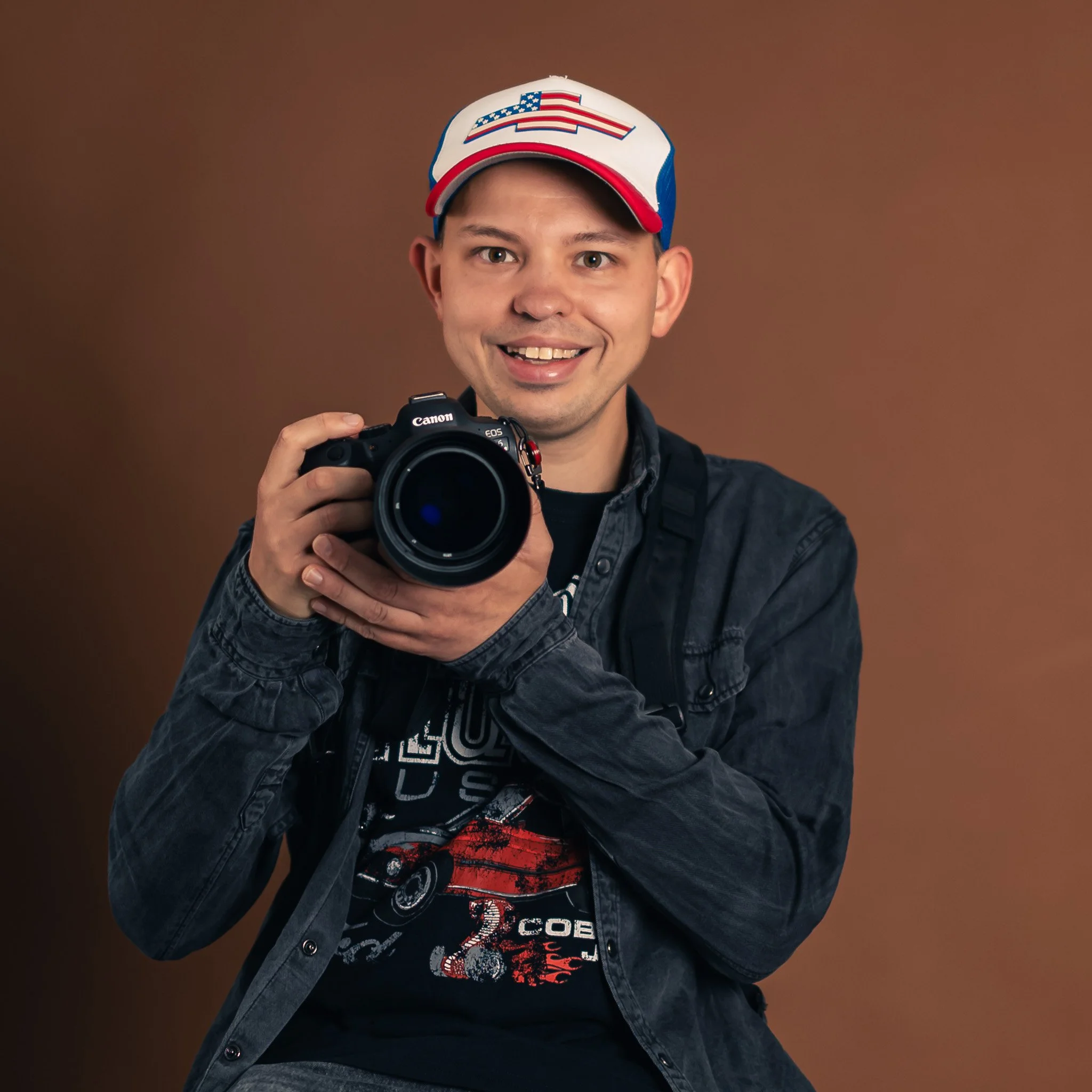 Roy de Rooij, photographer, holding a Canon camera, dressed in a black graphic t-shirt and denim jacket, posing against a brown background.
