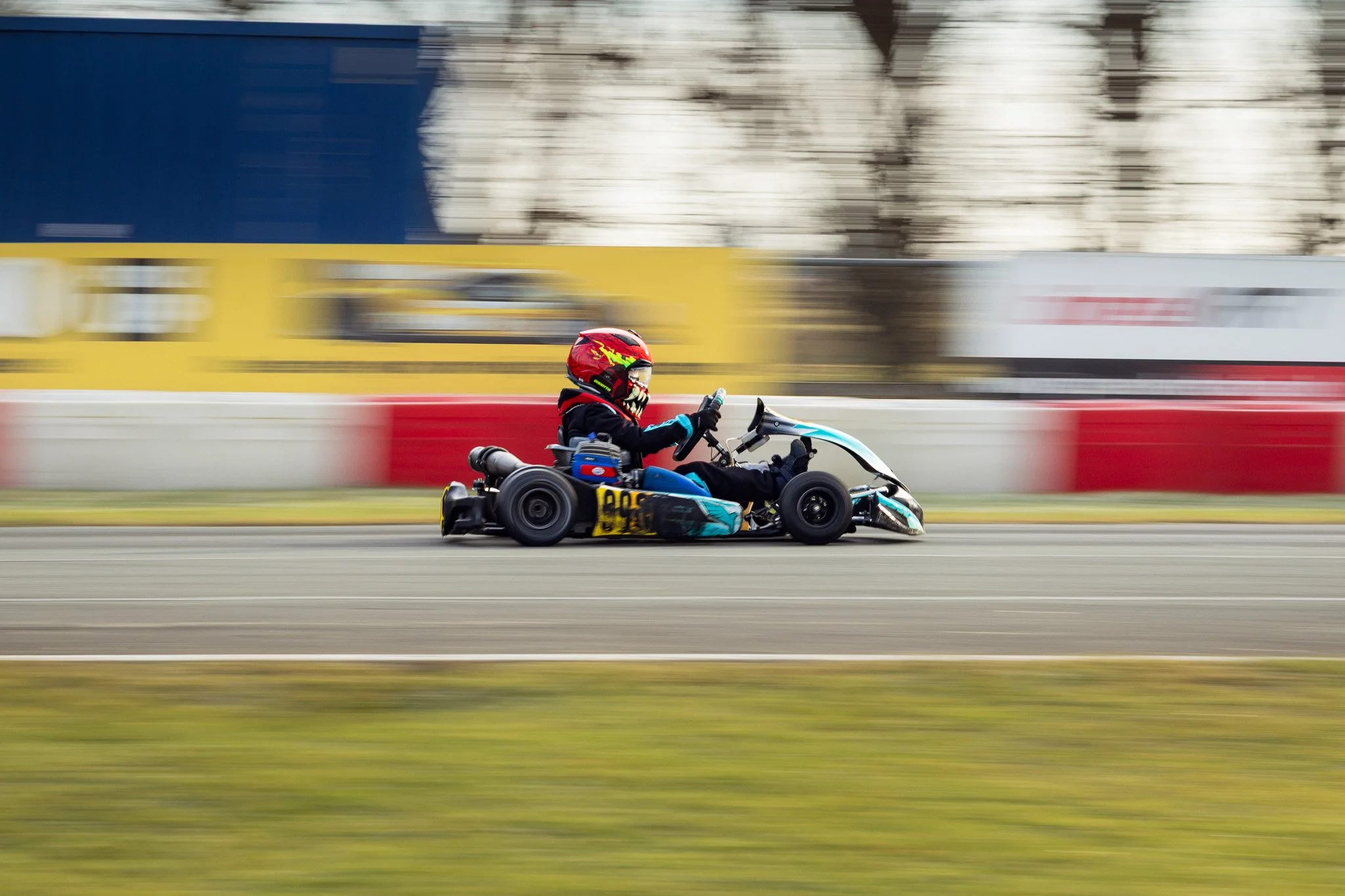 A child dressed in racing gear, including a helmet, riding a go-kart on a race track with blurred background and foreground, indicating high speed.