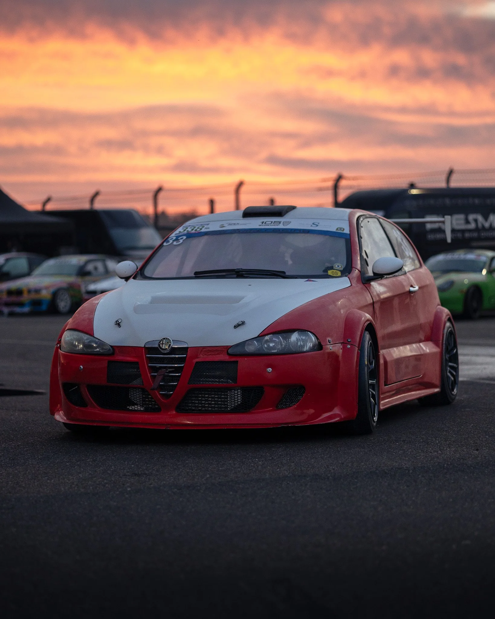 A red and white modified Alfa Romeo race car on a race track during sunset, with other race cars and trailers in the background.