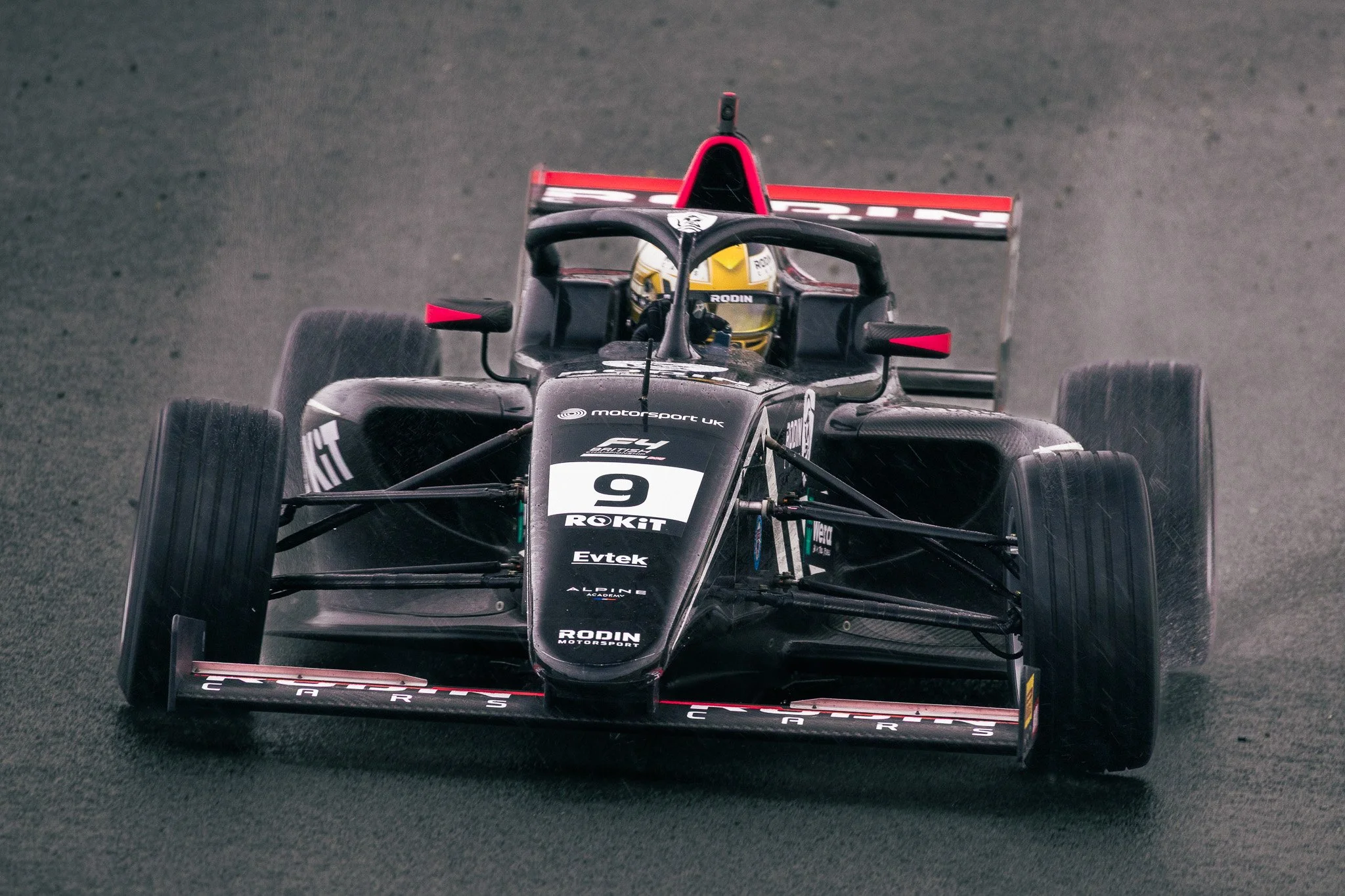 A black Formula race car with red accents speeding on a track, driven by a driver in a yellow helmet.