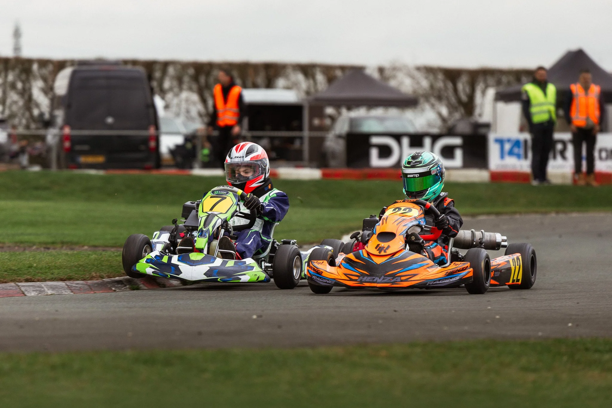 Two young go-kart racers in racing suits and helmets compete on a track, with officials standing in the background.
