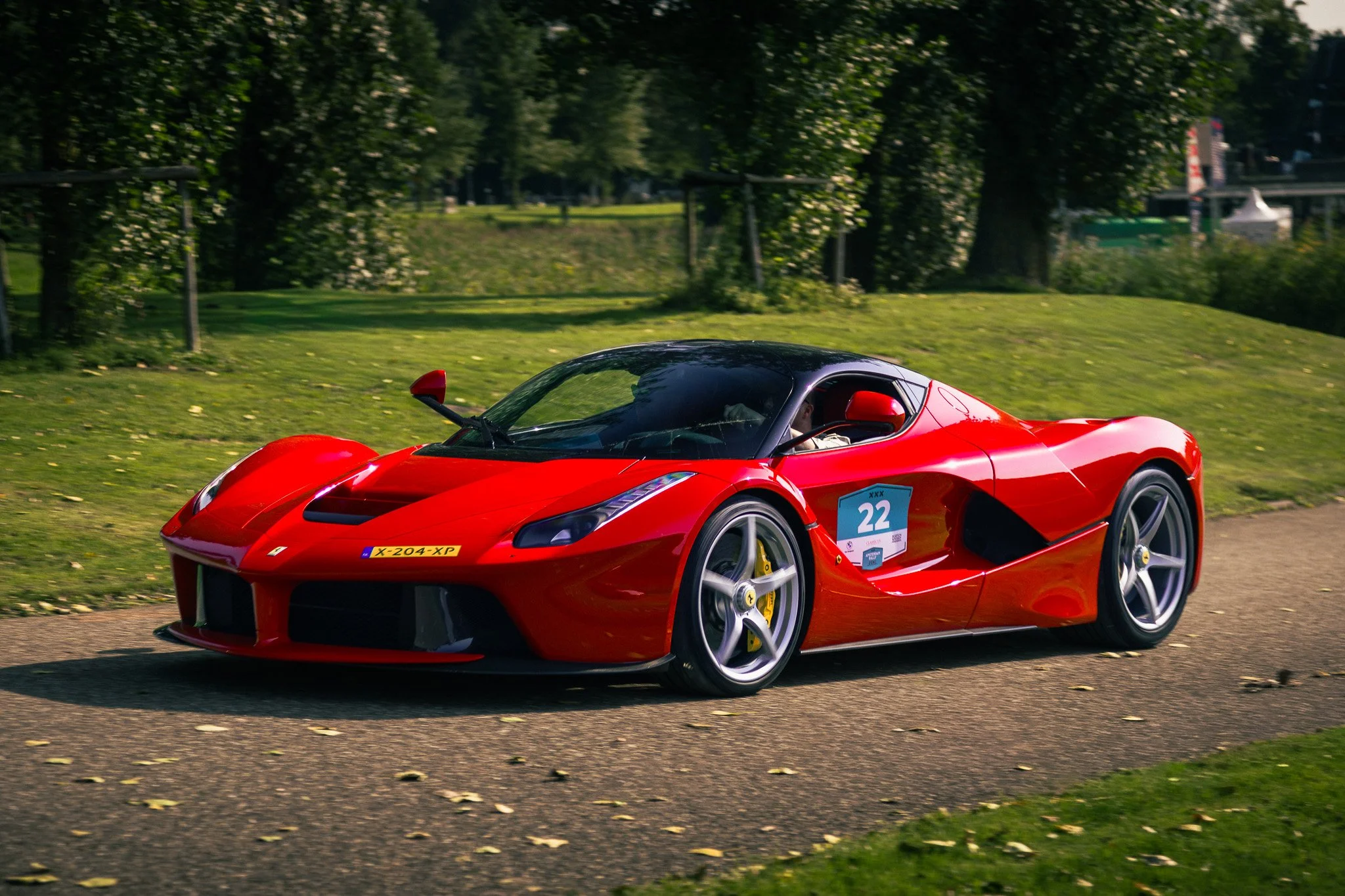 Red sports car driving on a paved road with green grass and trees in the background.