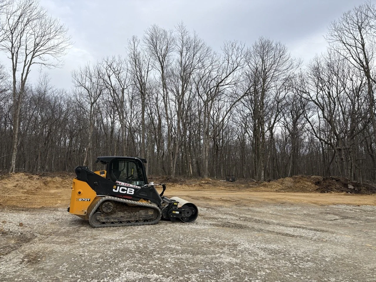 A small JCB tracked compact excavator on a construction site with bare trees in the background under a cloudy sky.