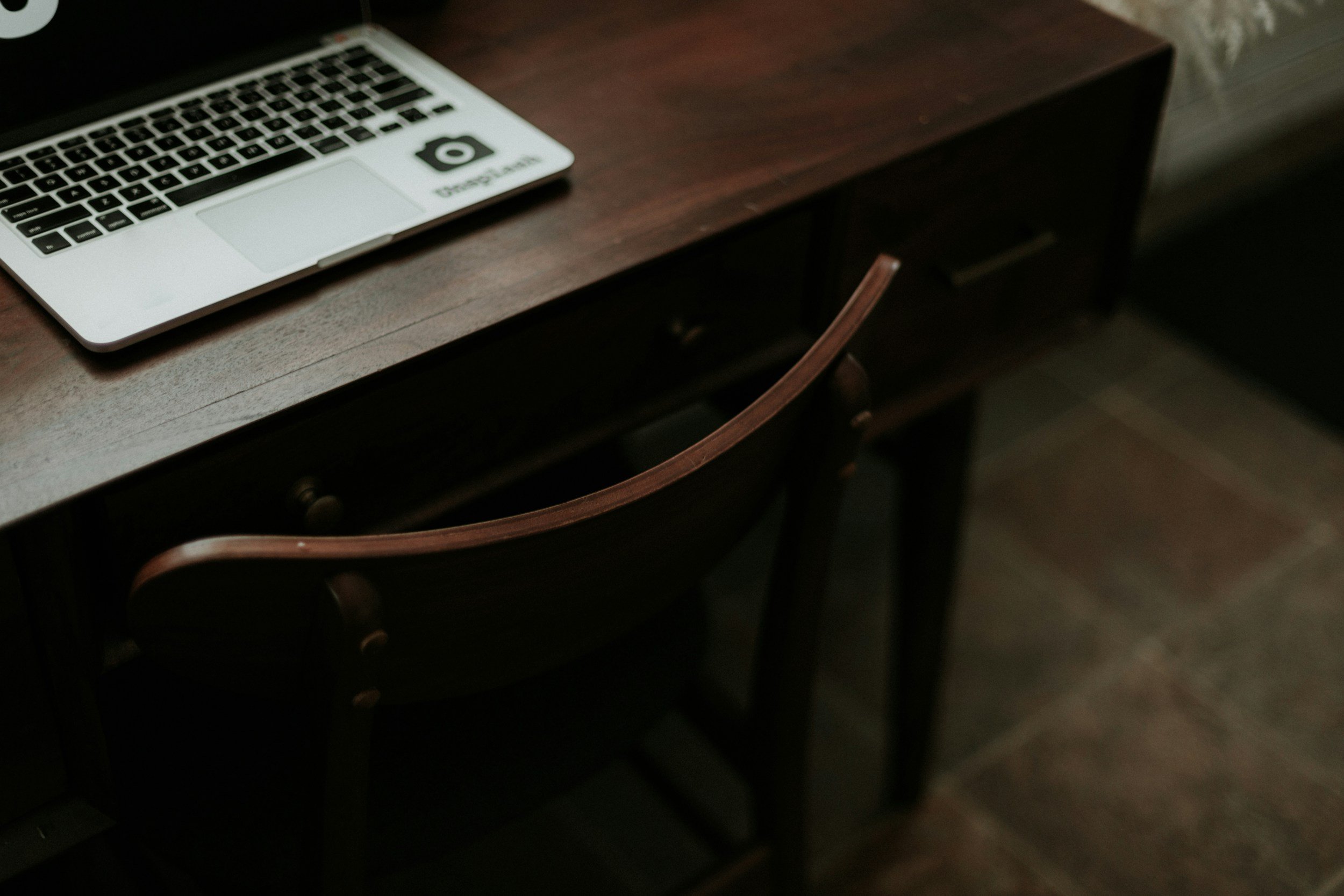 A laptop open on a warm, dark wood desk with an empty wooden chair, inviting authors into our distraction-free co-writing sessions.