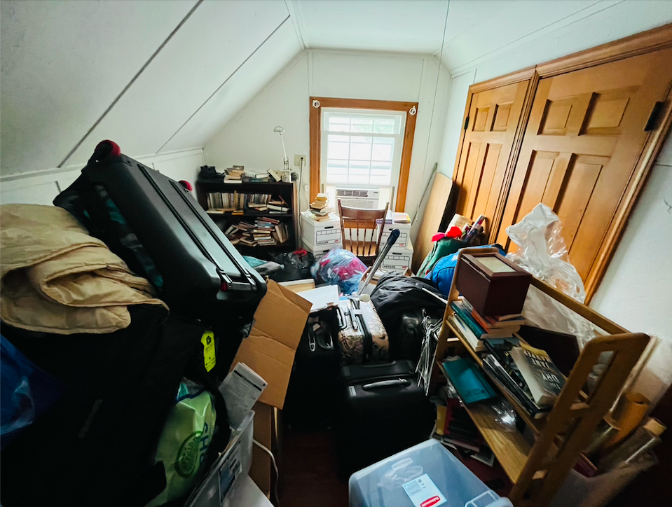 Cluttered room filled with suitcases, cardboard boxes, books, and various household items, with a small window at the far end.
