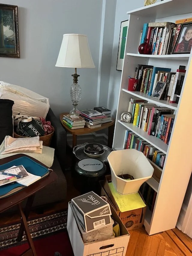 A cluttered living room corner with a white bookshelf filled with books, an adjacent table with a lamp and more books, assorted storage boxes, and miscellaneous items including a basket, fabric, and a tray on a chair.