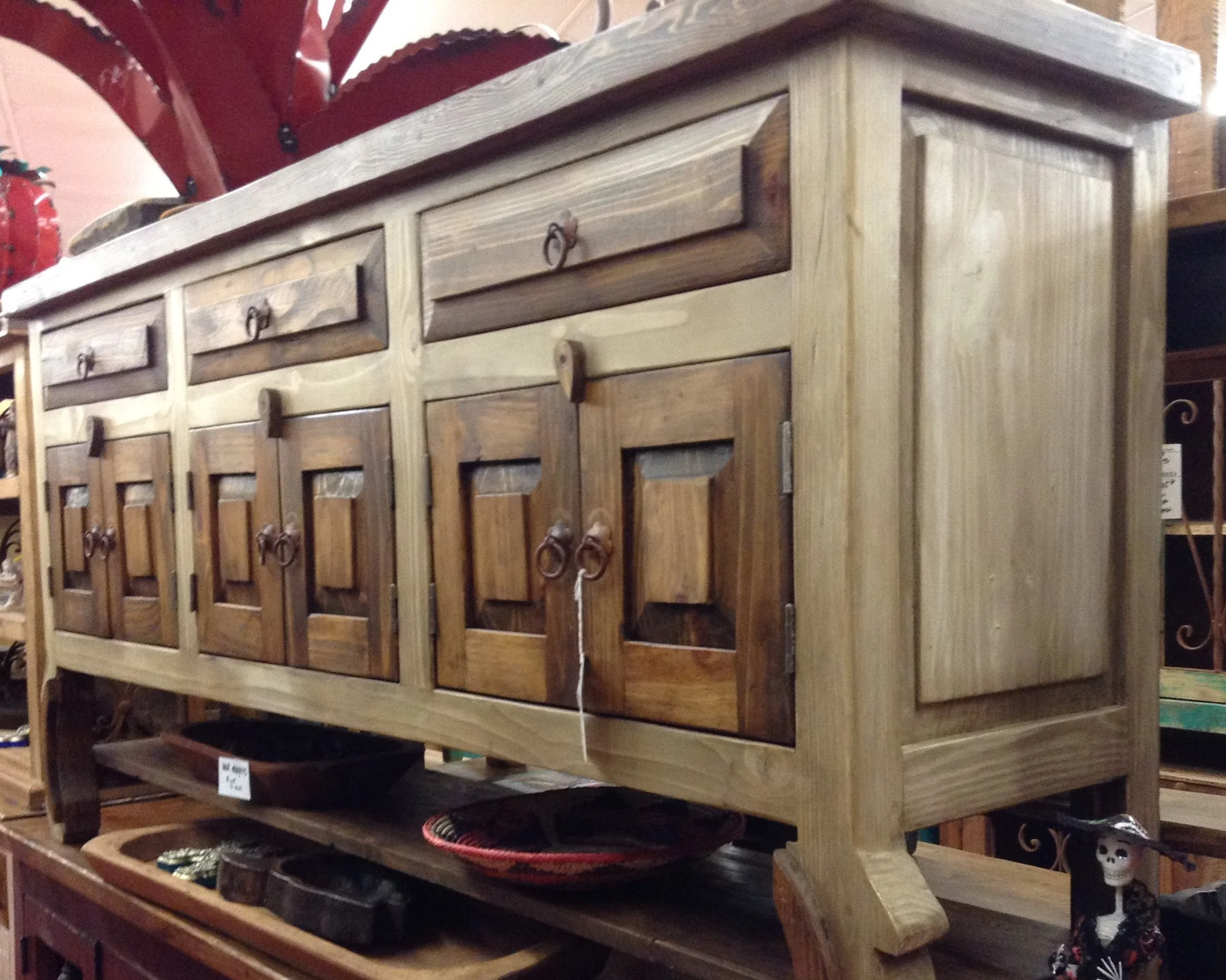 A wooden sideboard with multiple drawers and cabinet doors, displayed in a store, with decorative handle hardware, and various items on nearby shelves.