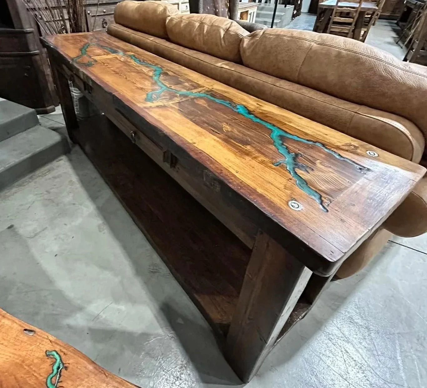 A rustic wooden table with a live edge and turquoise resin inlay next to a brown leather sofa, in a store with various furniture and wall art.