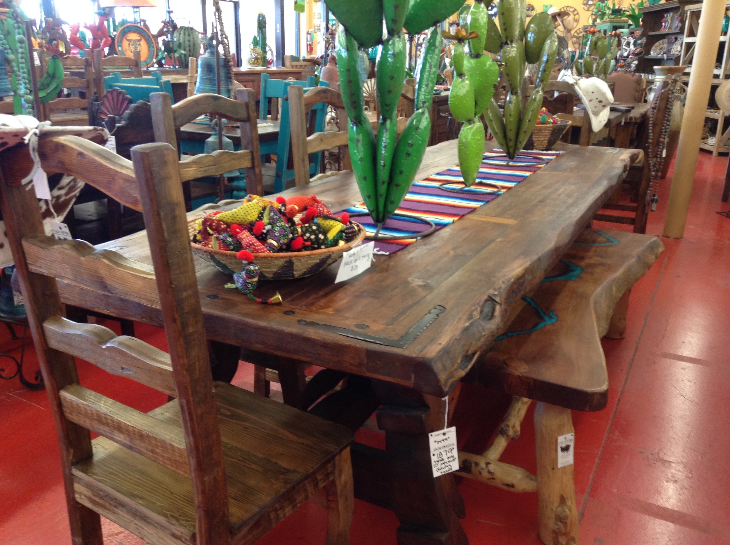 A rustic wooden dining table with a colorful woven table runner and a large cactus plant centerpiece. There are wooden chairs around the table and decorative items in the background.