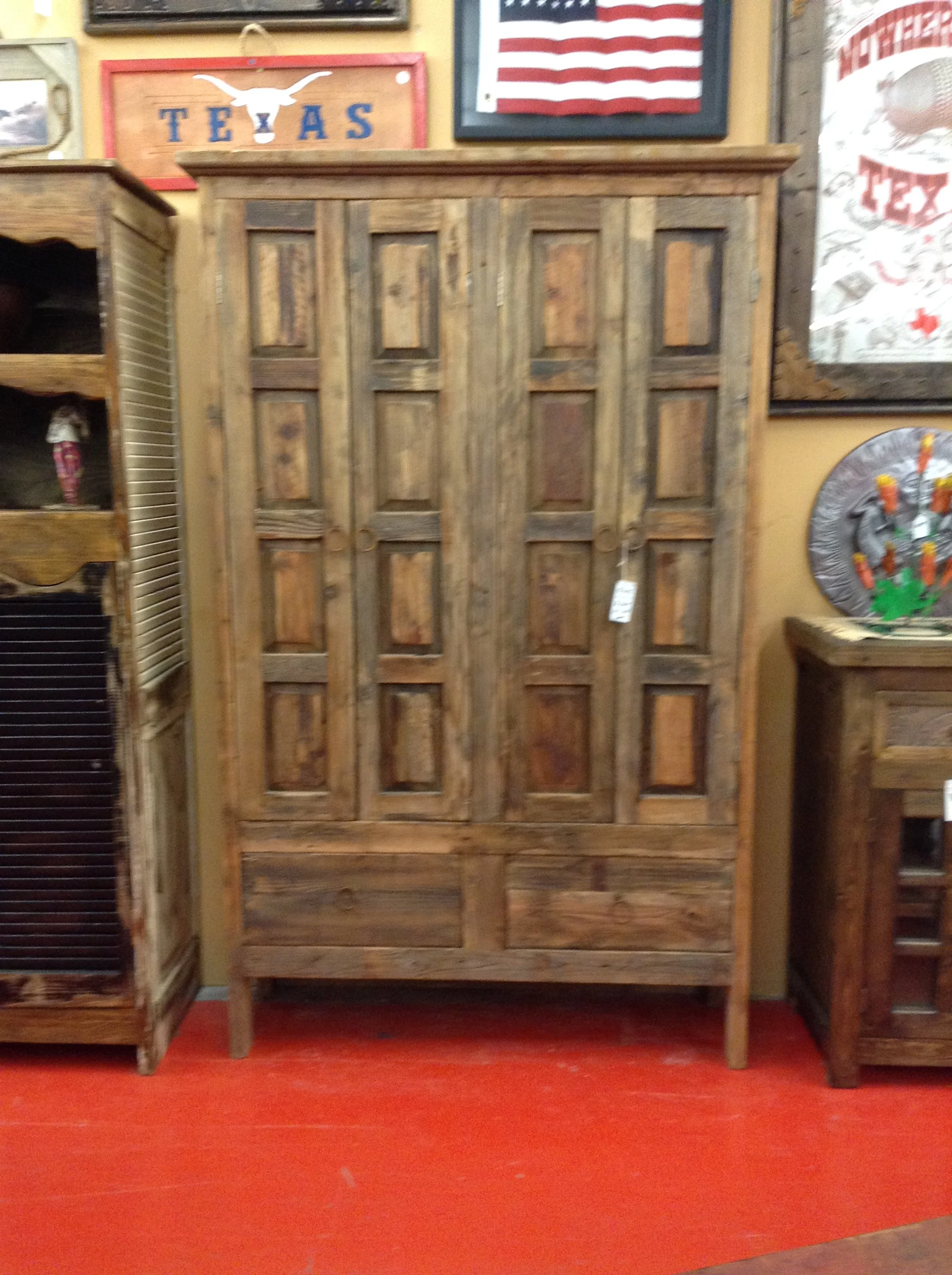 A wooden armoire with paneled doors in a rustic style, placed between two other wooden furniture pieces inside a store with various wall decorations including an American flag and Texas-themed signs.