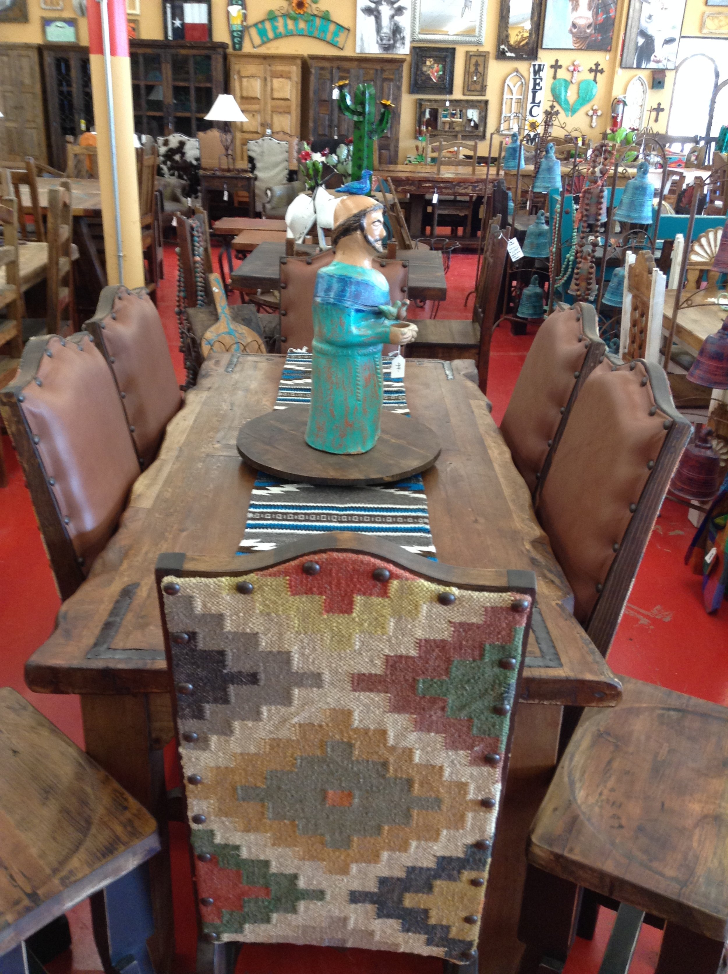 A rustic wooden dining table with eight chairs, six with leather upholstery and one with a colorful woven fabric backrest, is decorated with a blue and white striped table runner and a ceramic figurine of a bearded man in a blue robe at the center. The background features various wall art and decor, including a cactus sculpture and a sign that says "Hello".