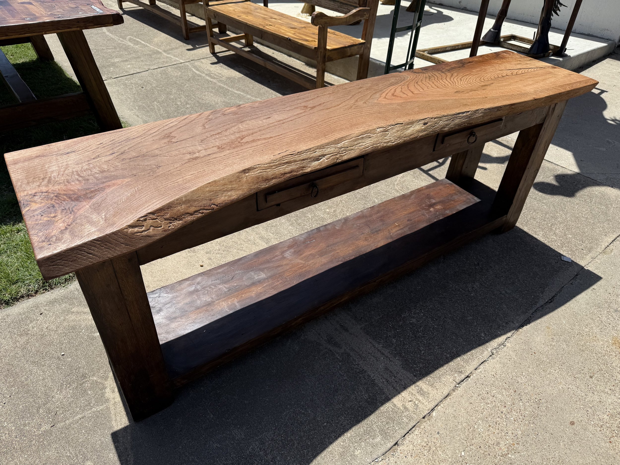 A rustic wooden table with a natural live edge, outdoor setting with other furniture and tools in the background.