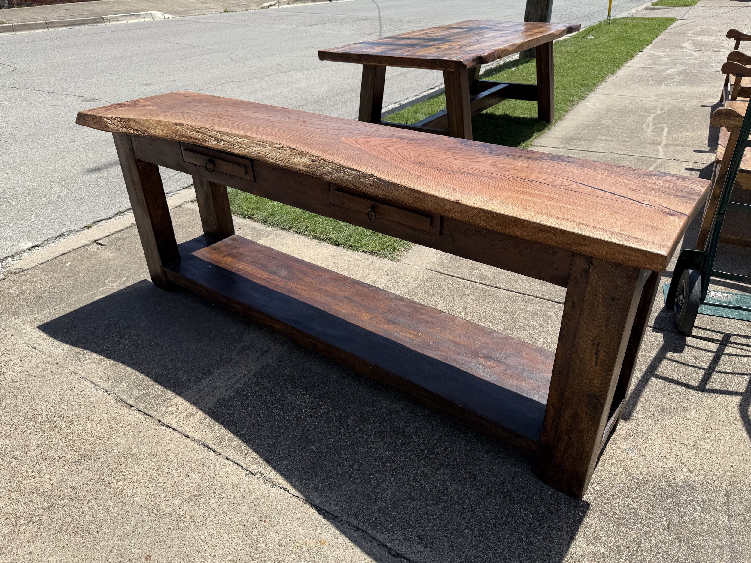 Outdoor wooden bench with a thick, polished wooden seat and a lower shelf, placed on a concrete sidewalk with grass and another wooden table in the background.