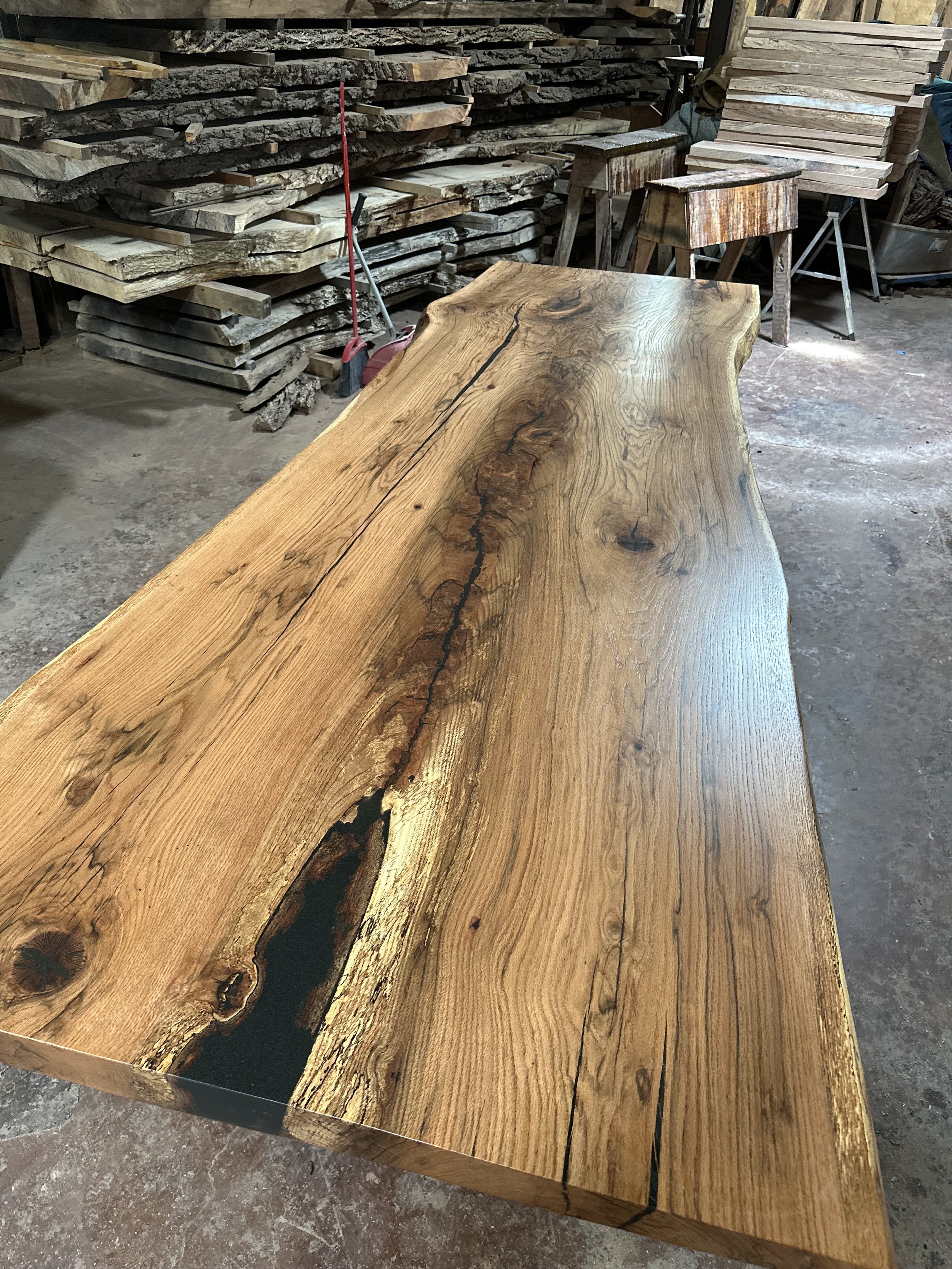 A large, polished wooden slab with natural wood grain and cracks, placed on a workshop floor with stacked wood planks and workbenches in the background.