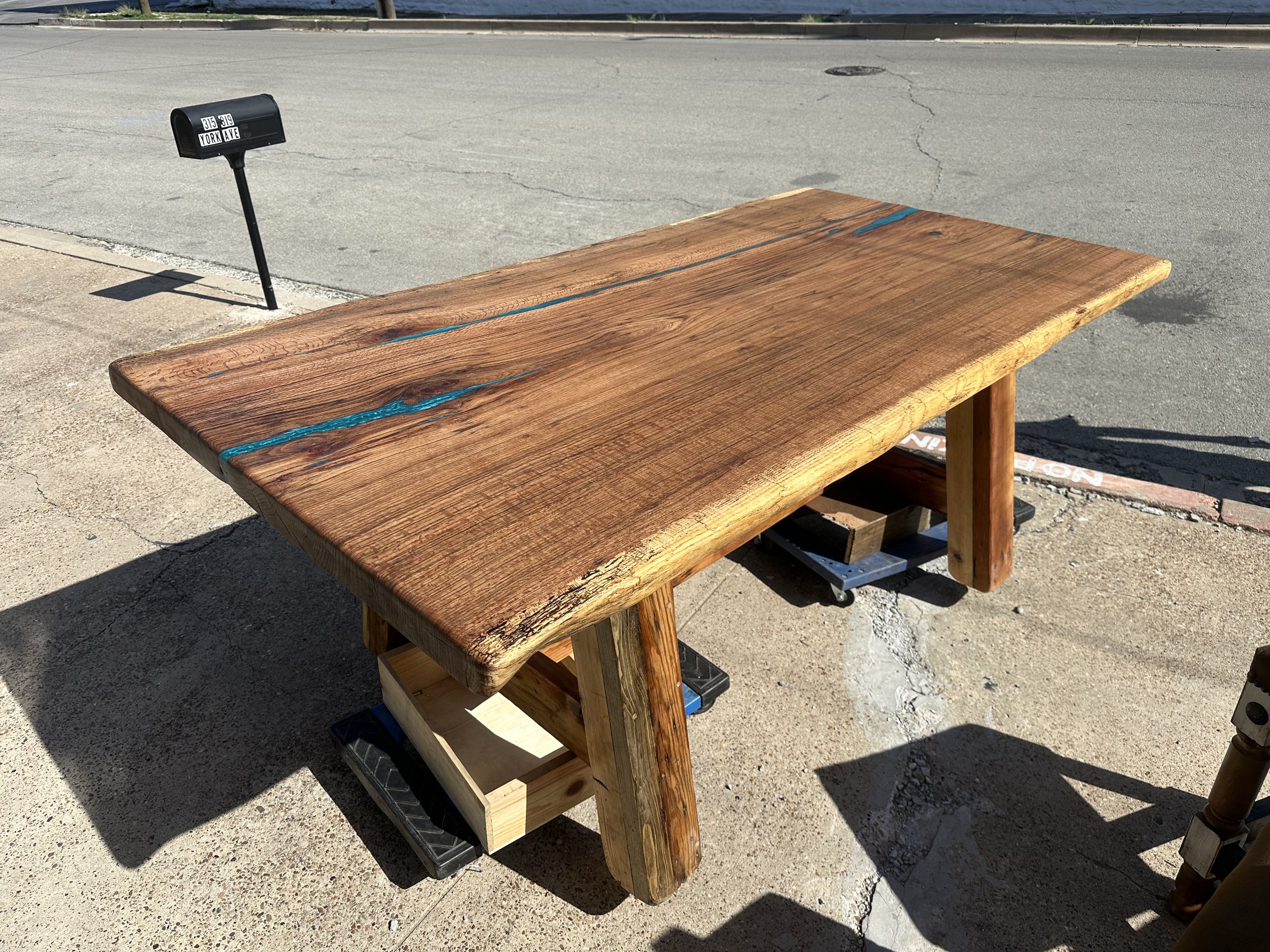 Handcrafted wooden table with a stained finish, placed outdoors on a sidewalk, with a small wooden box and a black object underneath.
