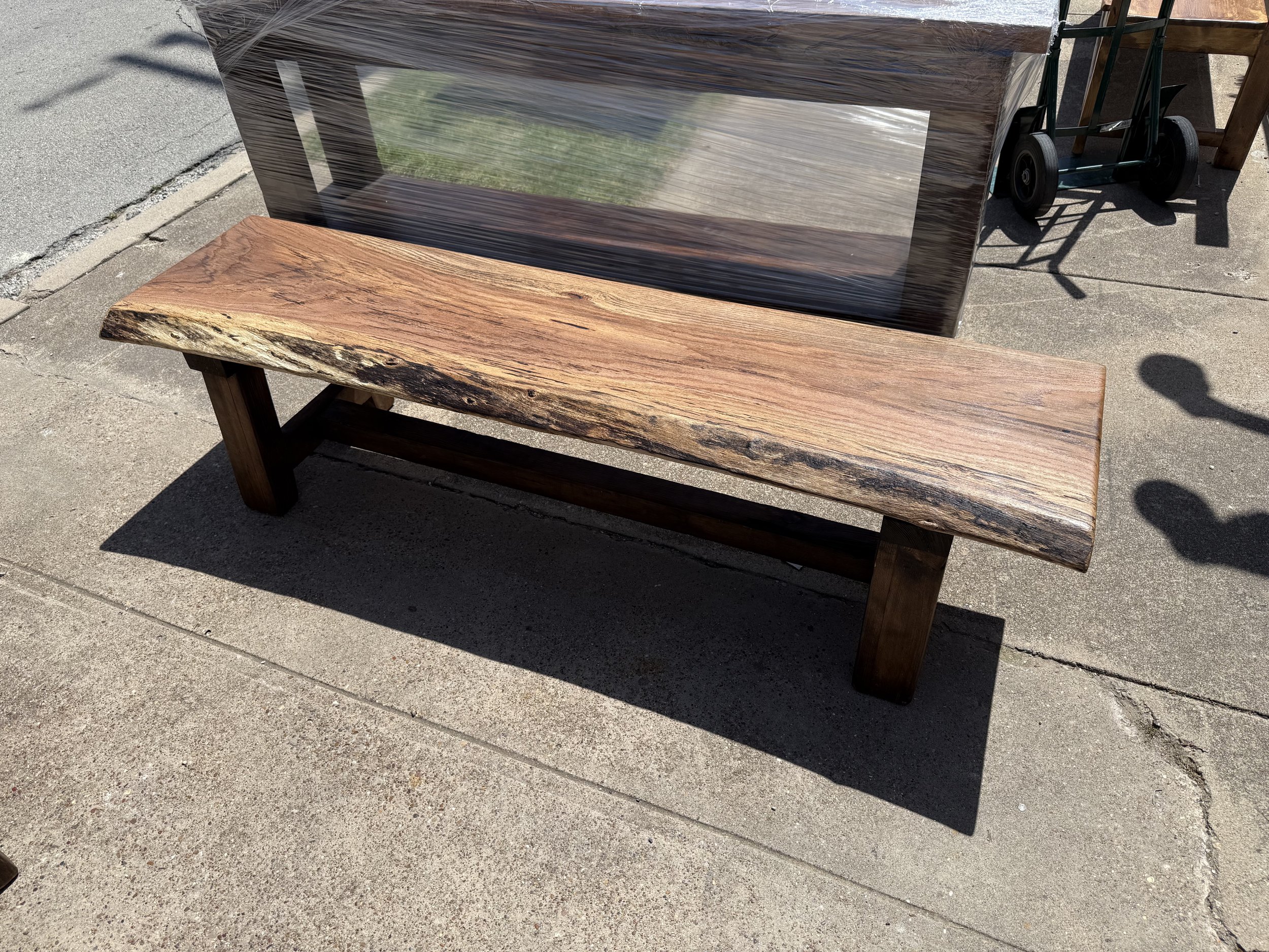 A wooden bench with a rustic, natural finish is placed on a concrete sidewalk outdoors. It has a simple design with two sturdy legs and a flat top. Behind the bench, there is a piece of furniture covered in plastic wrap, a small dolly, and a wooden t