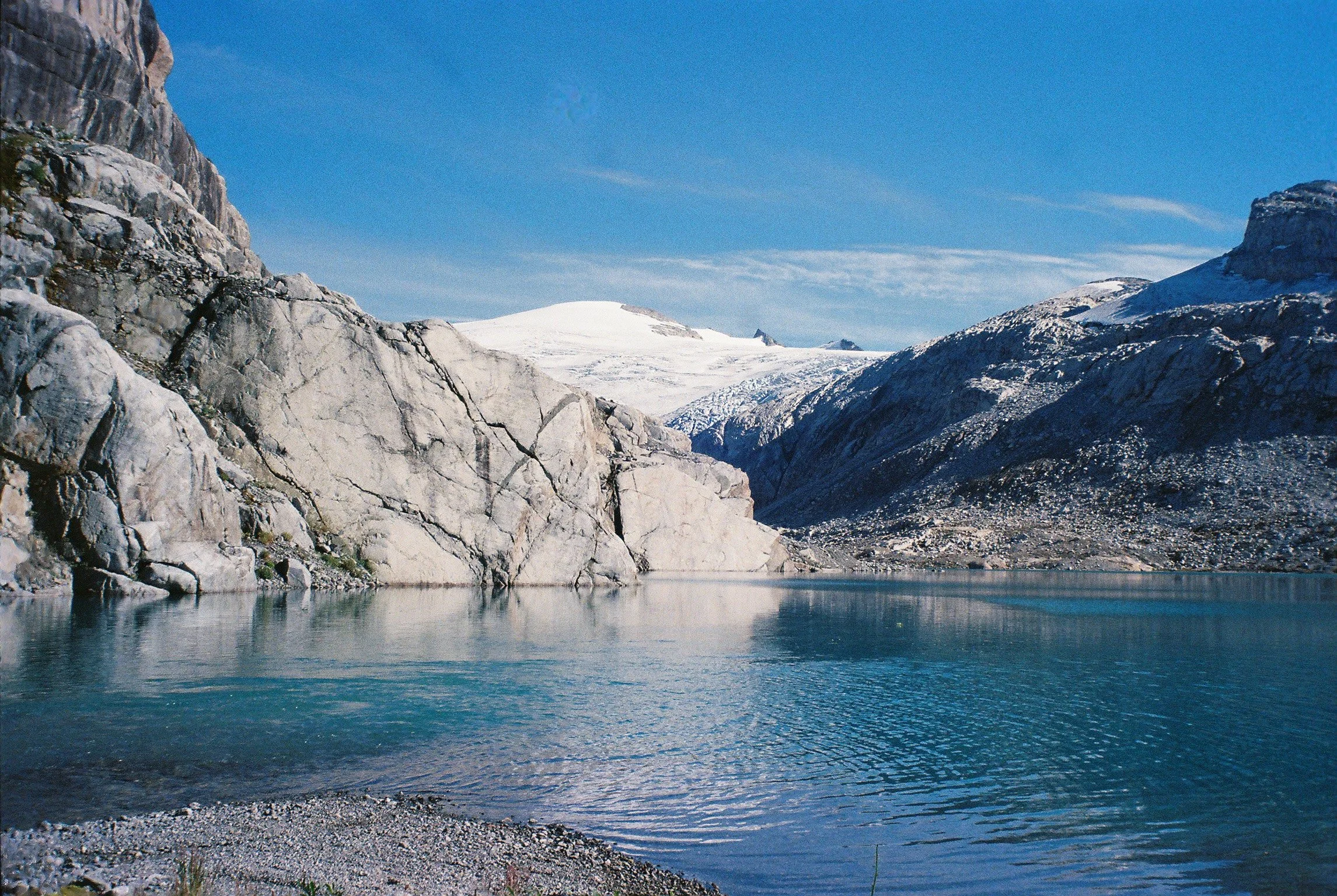 A scenic mountain landscape with a glacier and a clear blue lake in the foreground under a partly cloudy sky.
