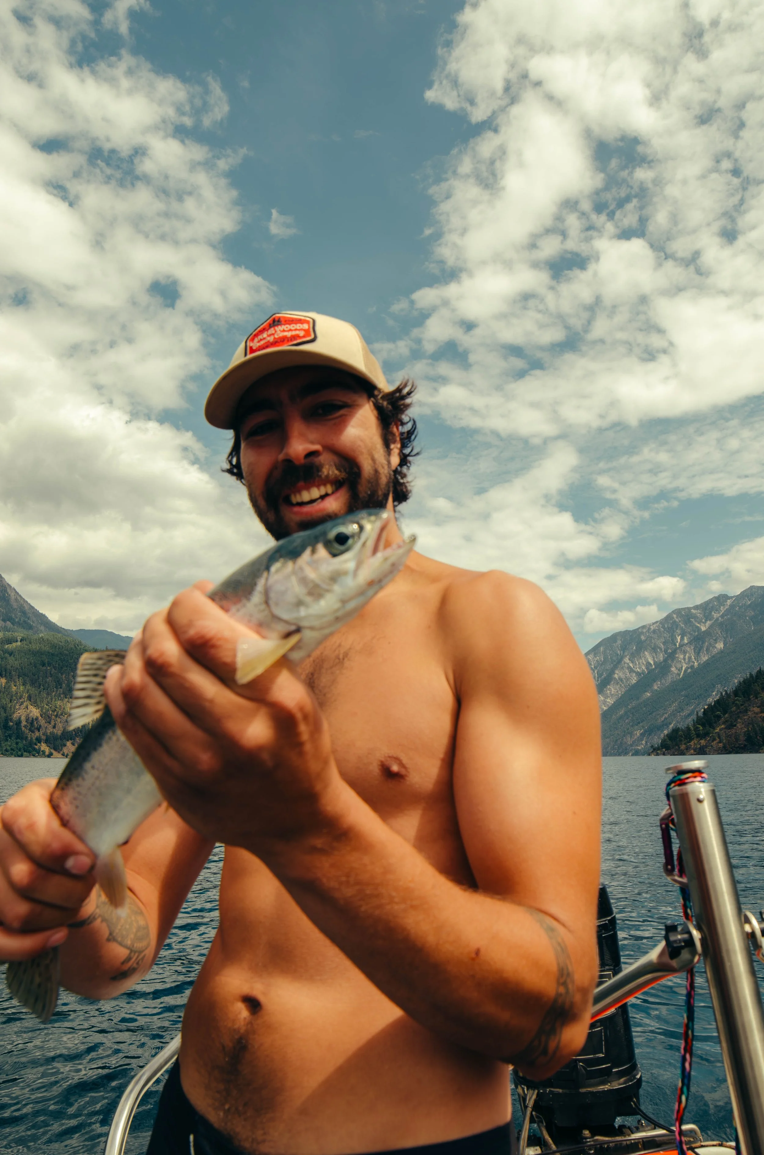 Smiling shirtless man on a boat holding a freshly caught fish with mountainous landscape and cloudy sky in the background.