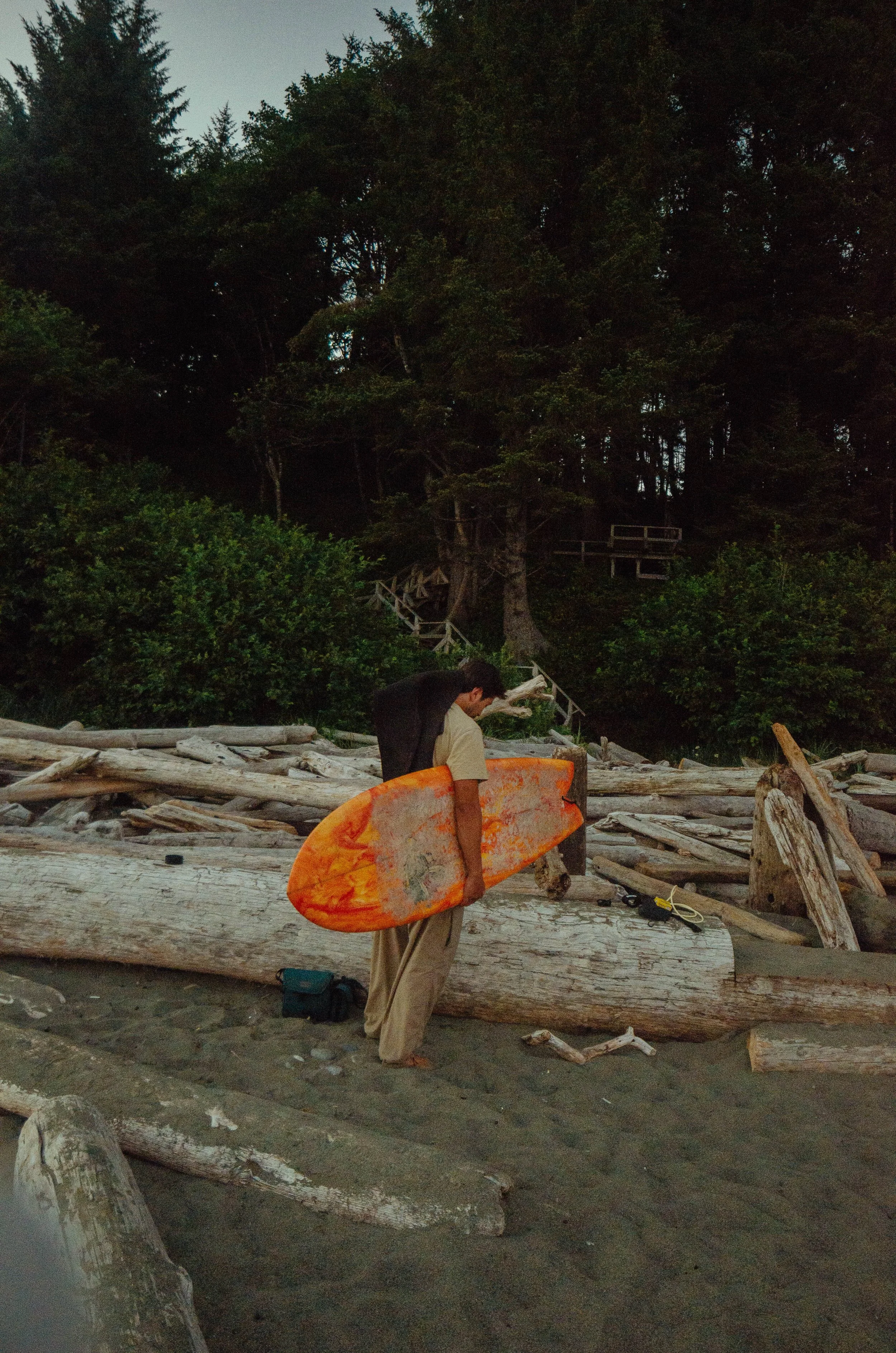 A person carrying an orange surfboard on a beach with driftwood and trees in the background.