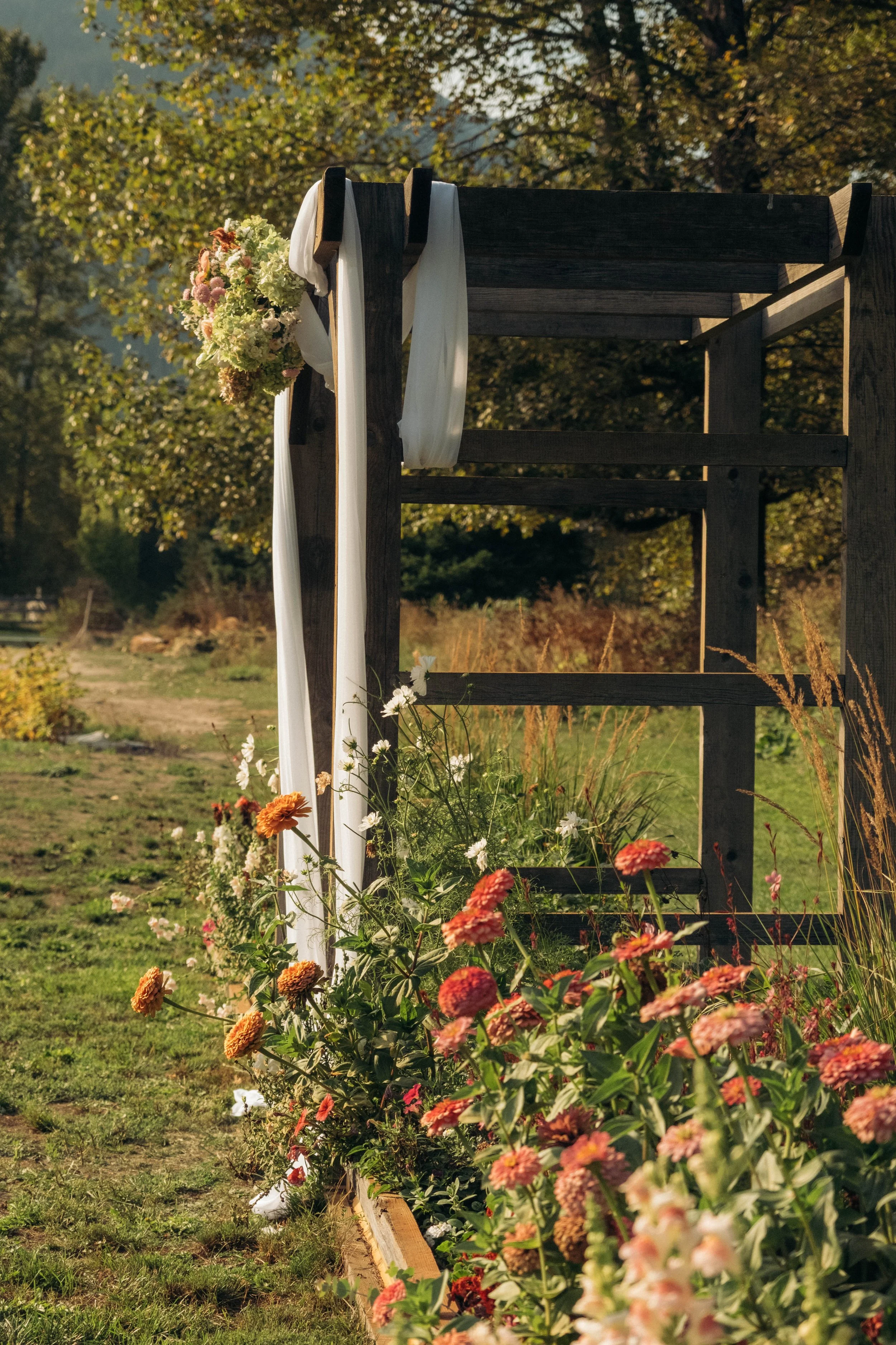 Wooden wedding arch decorated with white and pink flowers, draped with white fabric, set in a garden with colorful flowers and greenery in the background.