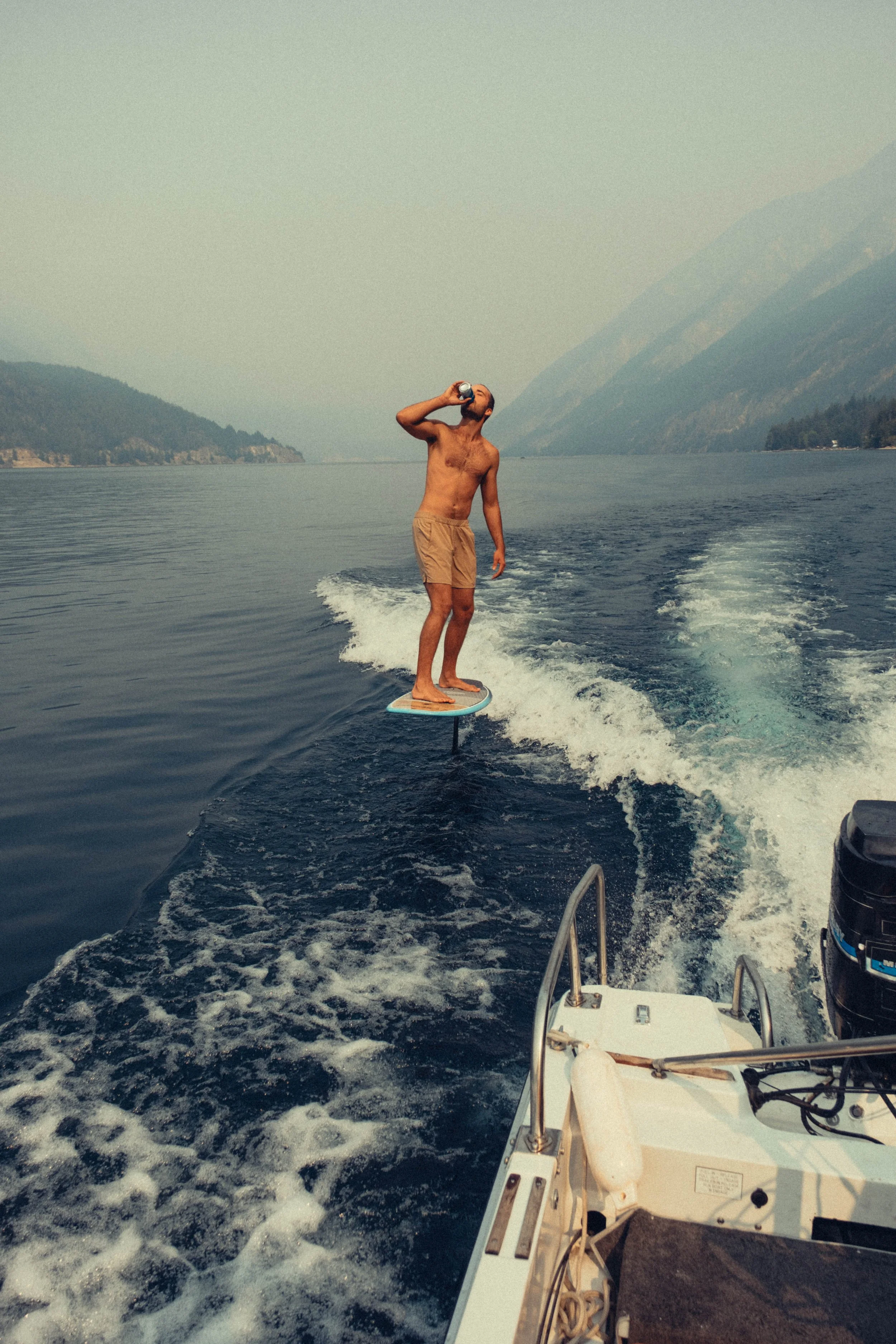 A man standing on a wakeboard is being pulled by a boat on a body of water, drinking from a can, with mountains in the background.