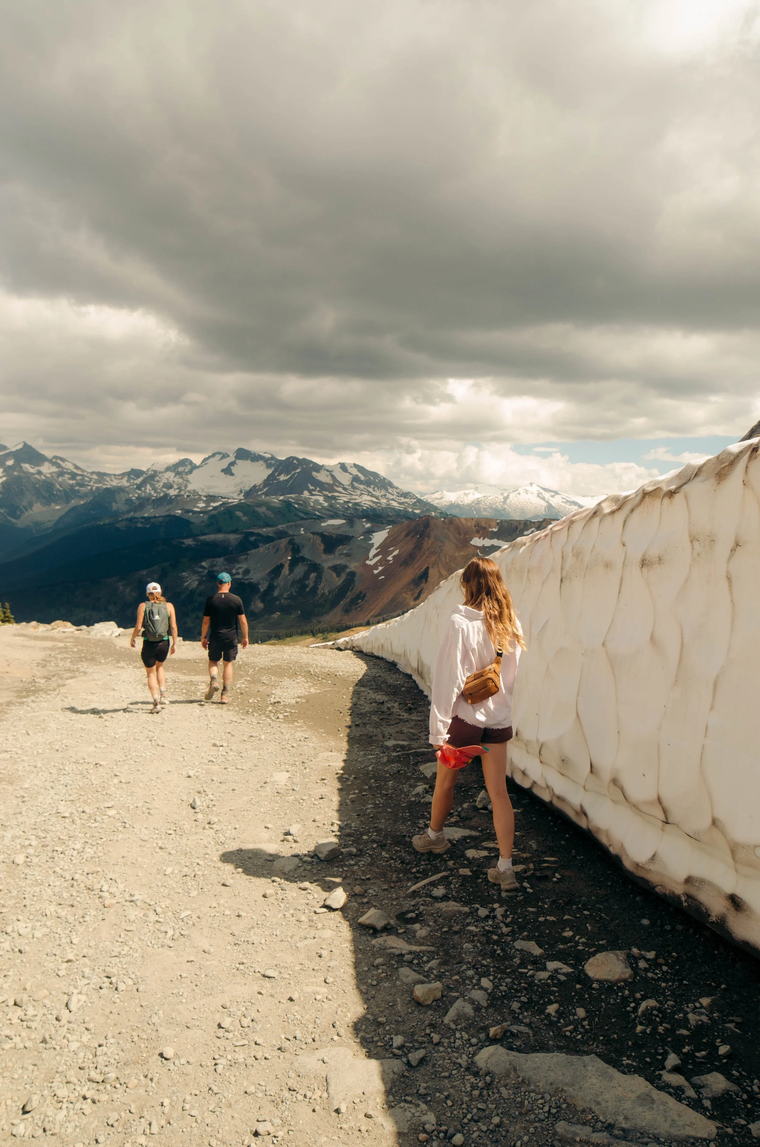 Three hikers walking along a mountain trail beside a wall of snow with snow-capped peaks in the background and cloudy sky.