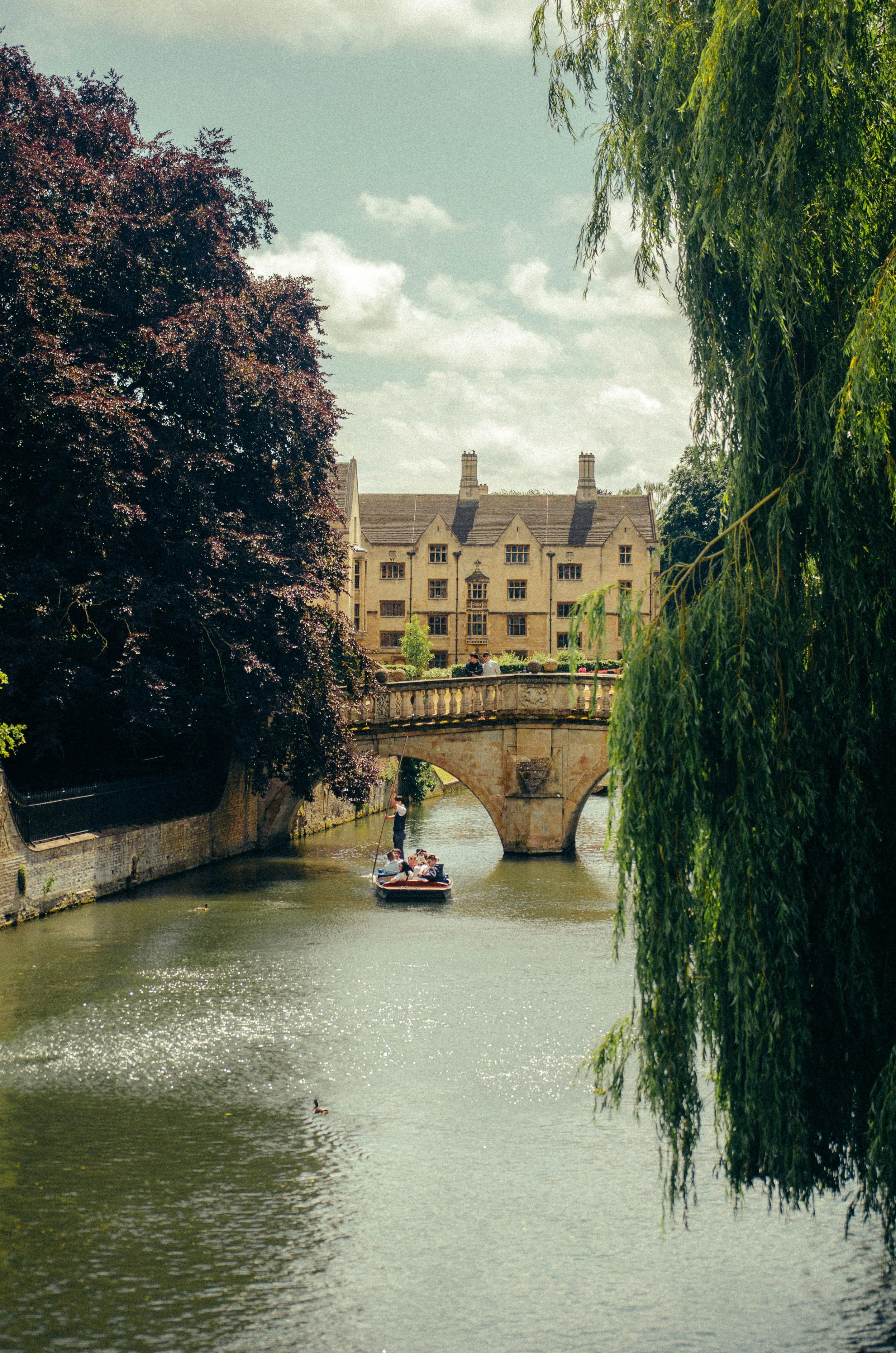 A picturesque river scene with a stone bridge, a boat with tourists, and historic buildings in the background. Tall trees line both sides of the river.