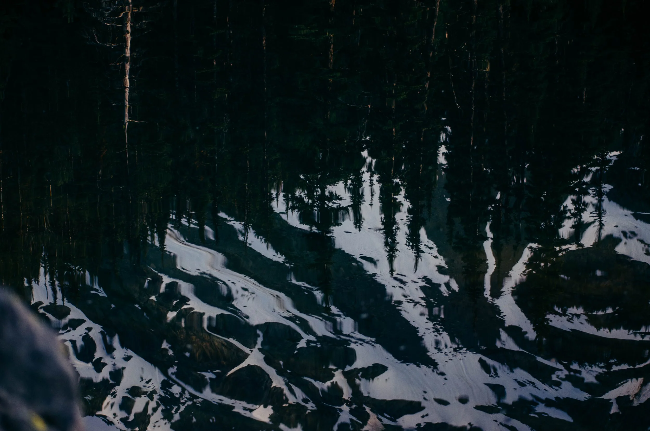Reflection of evergreen trees and snowy mountain peaks on a mountain lake with rocks in the foreground.