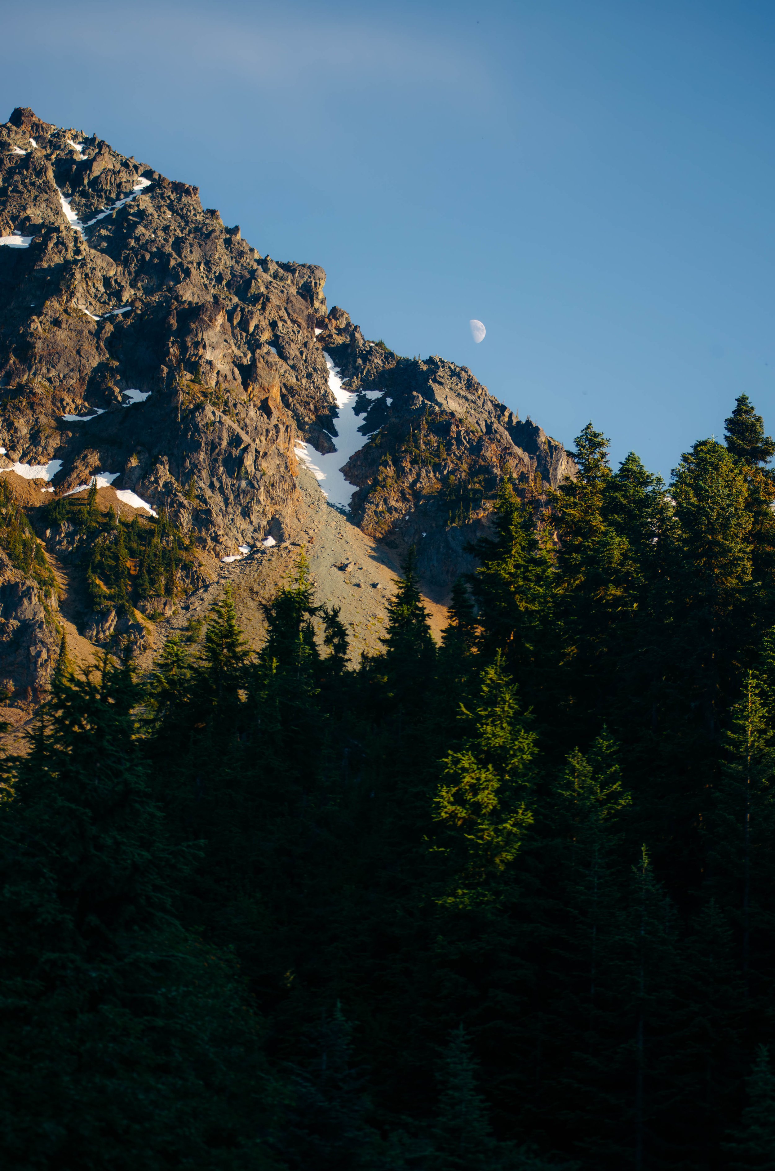A mountain landscape with rocky peaks, snow patches, green pine trees, and a visible moon in the clear blue sky.