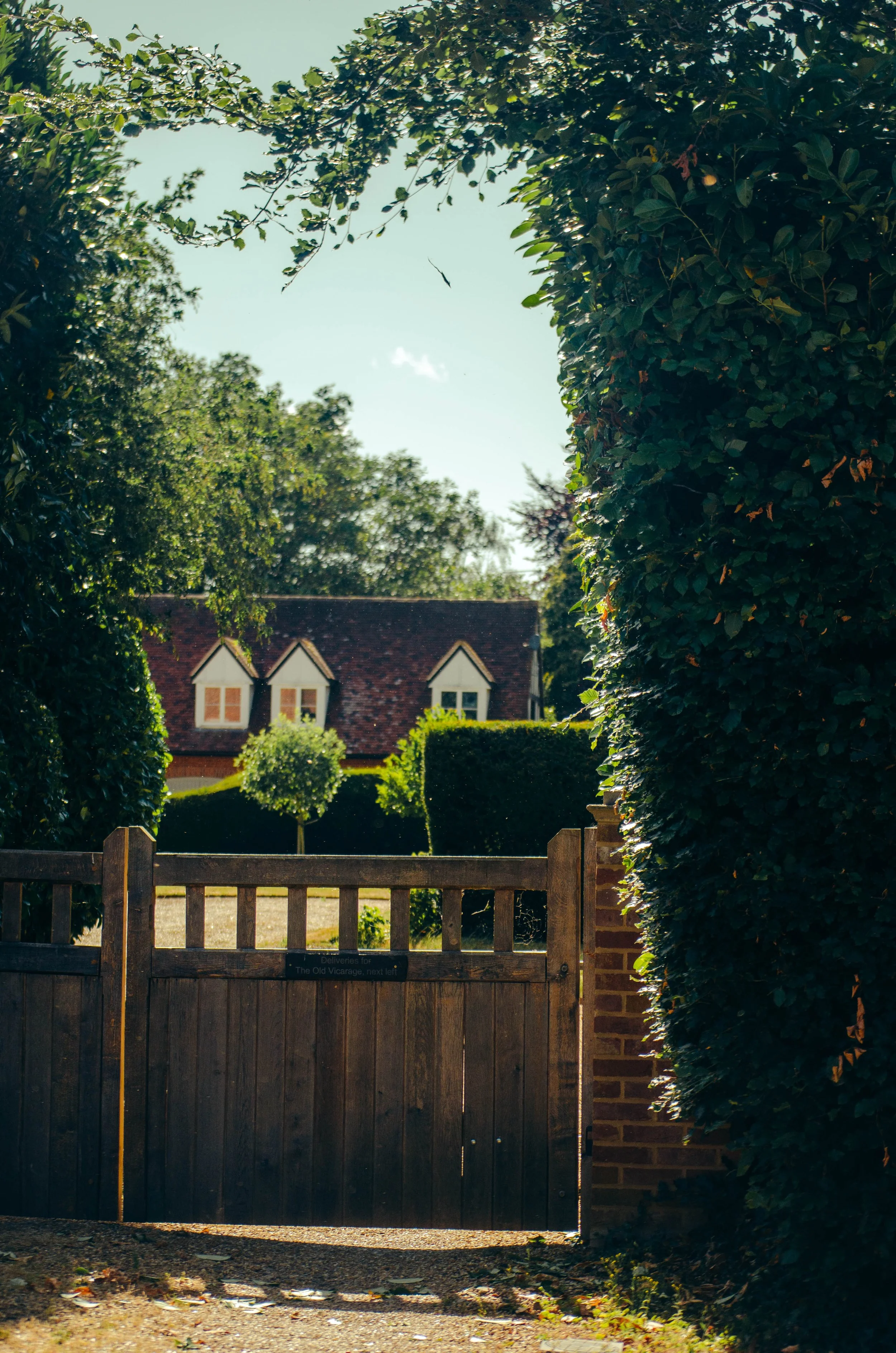A wooden gate in front of a garden with neatly trimmed hedges and a small tree, with a house with a red roof and dormer windows in the background, surrounded by lush green trees.
