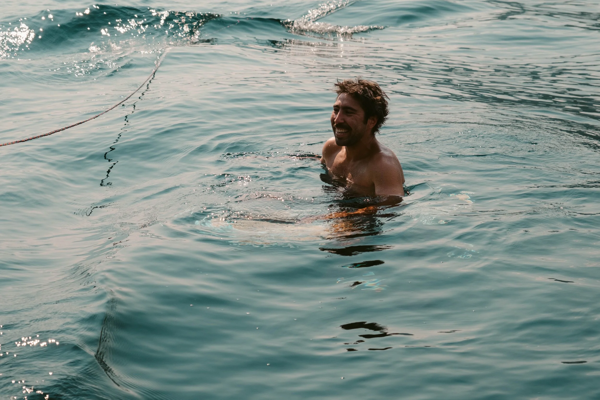 A man with dark hair and a beard swimming in the water, smiling and looking to the side, with a rope floating nearby in the water.