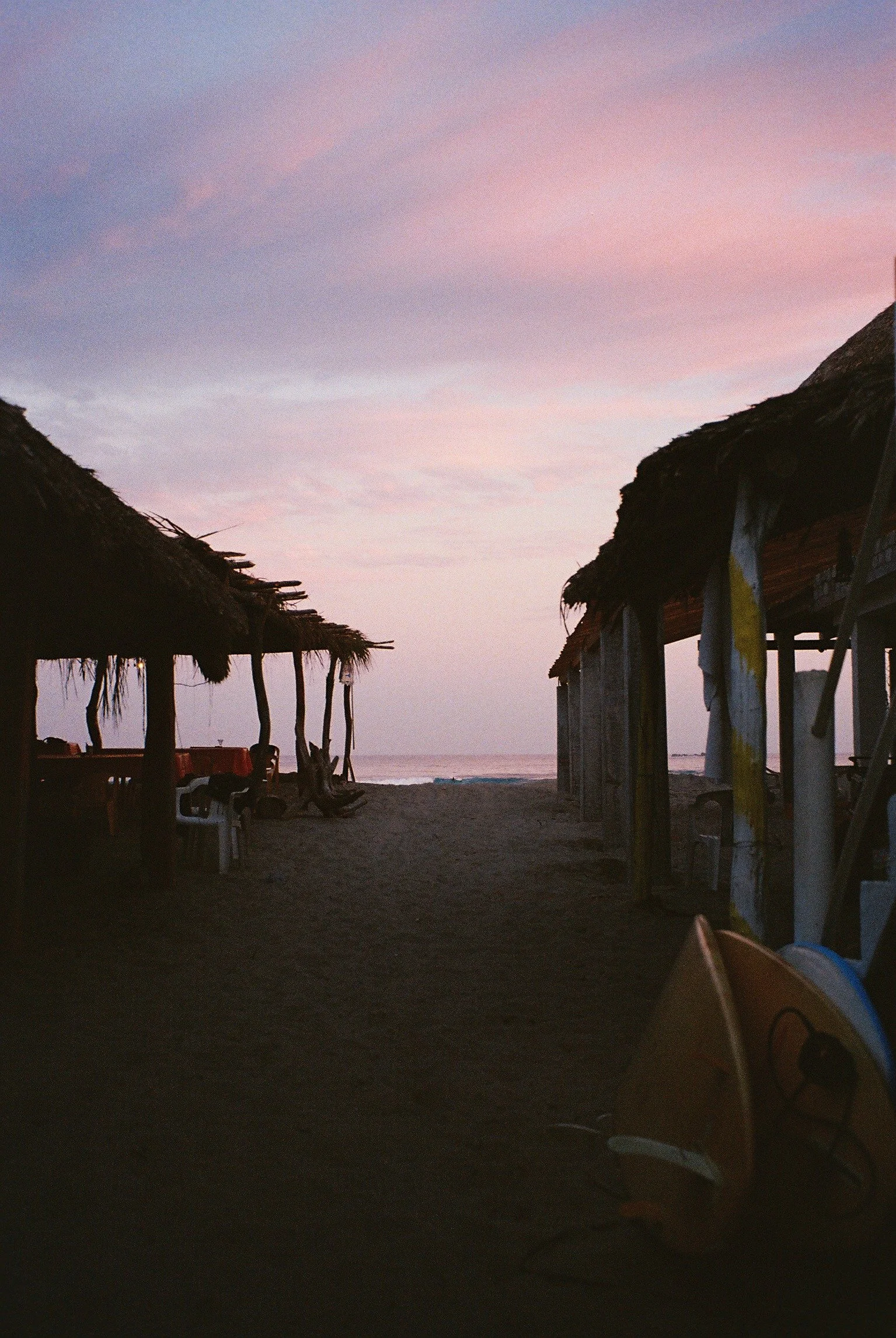Beach huts with thatched roofs at sunset on a sandy beach, with a kayak visible in the foreground.