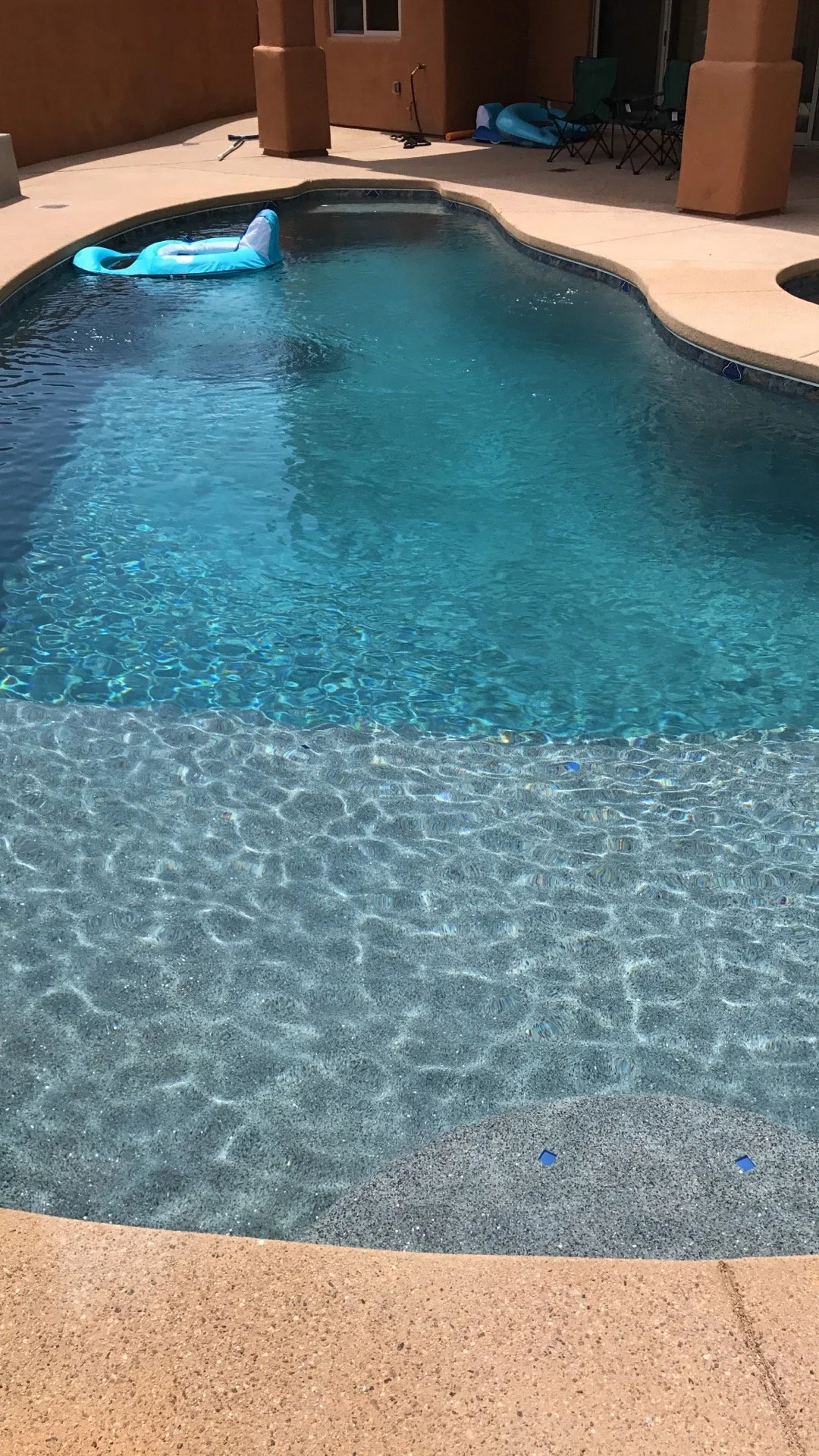 A backyard swimming pool with clear blue water and an inflatable float, surrounded by a beige concrete deck and patio furniture in the background.