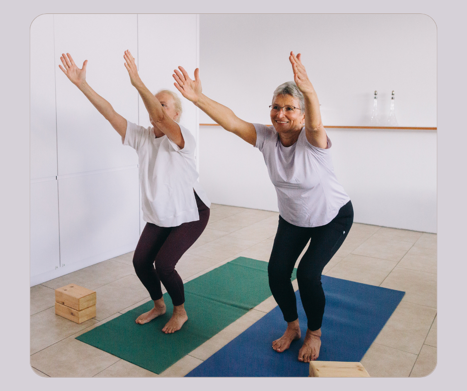 Two middle aged women practicing yoga, standing on yoga mats with arms raised, smiling, indoors with a white wall background.