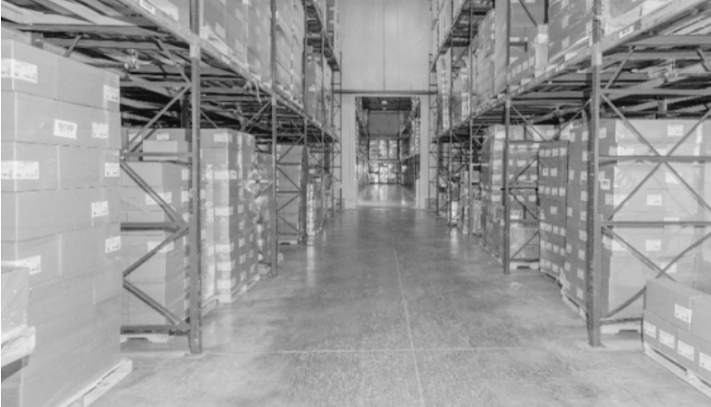 Warehouse interior with rows of stacked boxes on metal racks, concrete floor, and high ceiling.