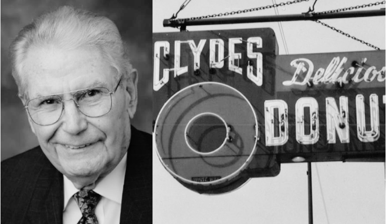 Black and white photo of an elderly man in glasses, wearing a suit, next to a vintage sign for Clyde's Delicious Donuts.