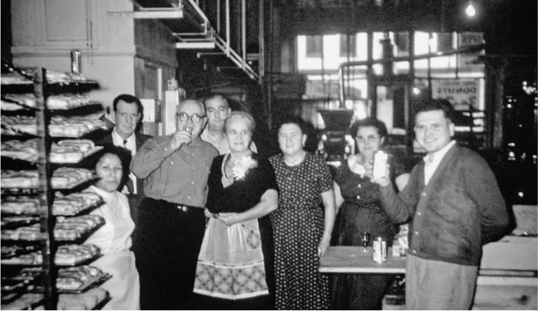 Vintage black and white photo of a group of people in a bakery, surrounded by trays of baked goods, smiling and holding drinks, with industrial equipment in the background.