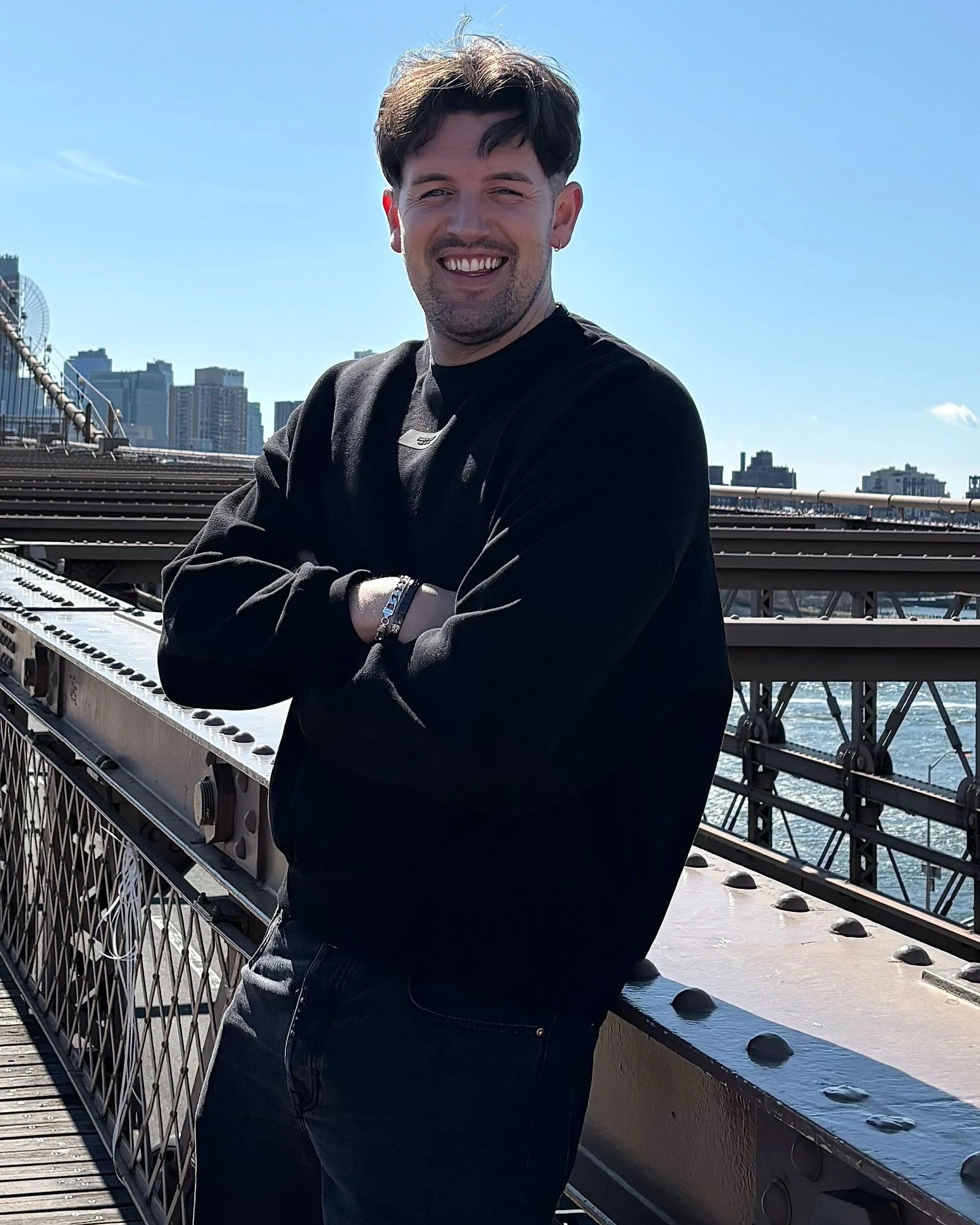 A smiling man with crossed arms standing on a bridge with city skyline and water in the background.