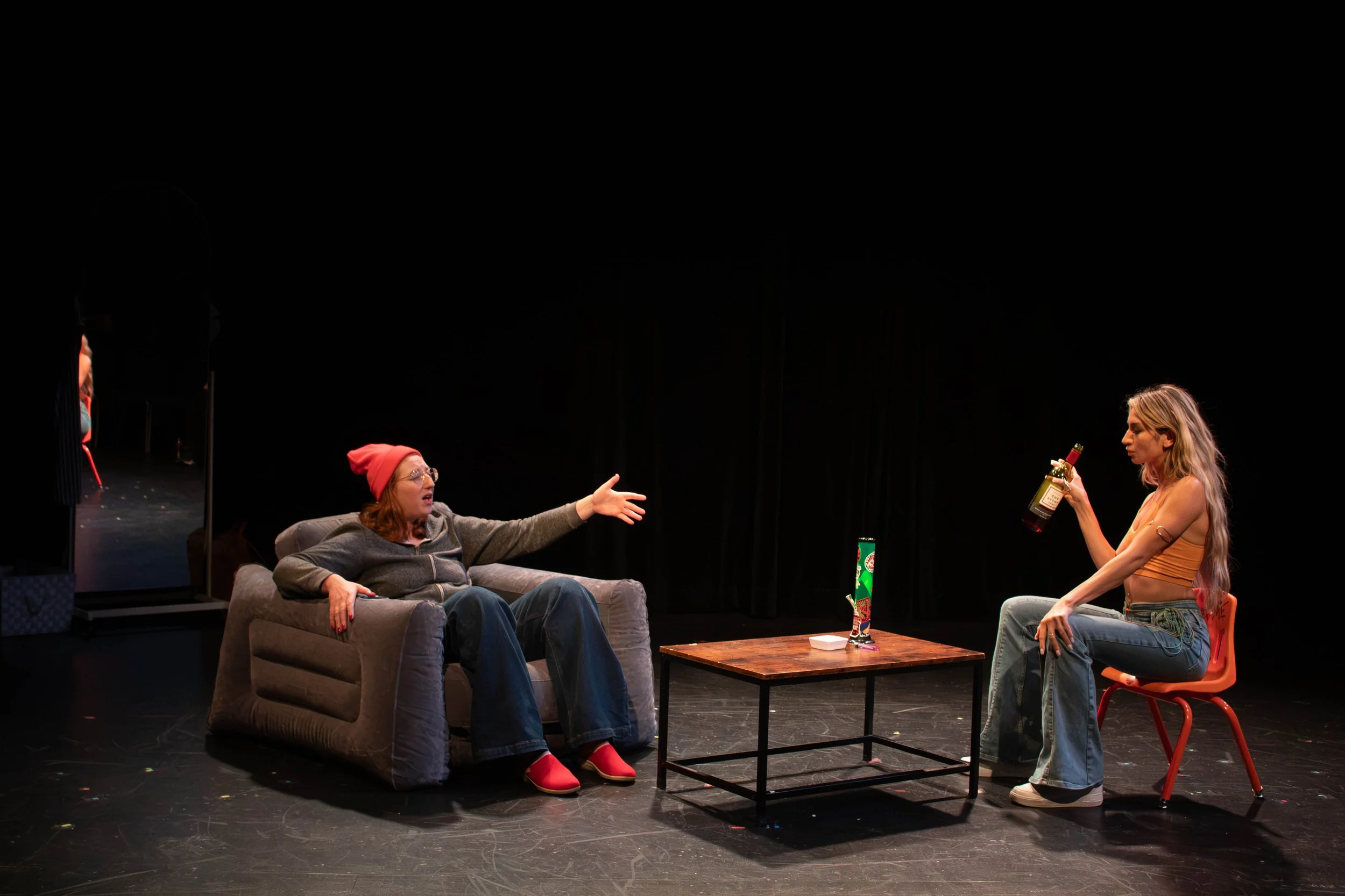 Performer Vanessa Meyer sitting in an inflatable chair gesturing during a stage production alongside a co-star holding a bottle.