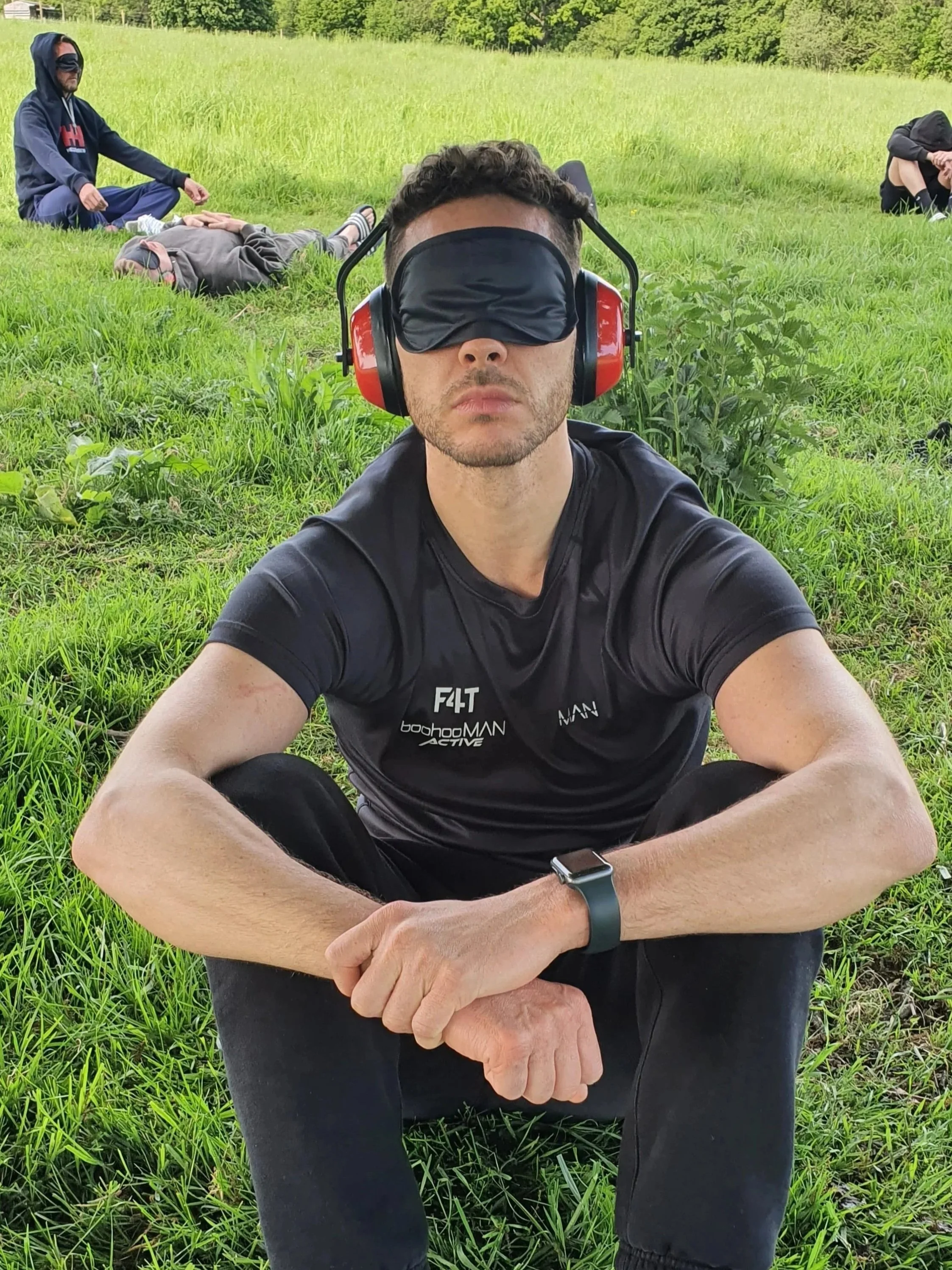 Man sitting outdoors on grass with his legs crossed, wearing a black T-shirt with logos, a smartwatch, and a blindfold, with headphones over the blindfold. In the background, several other people are lying or sitting on the grass in a field.