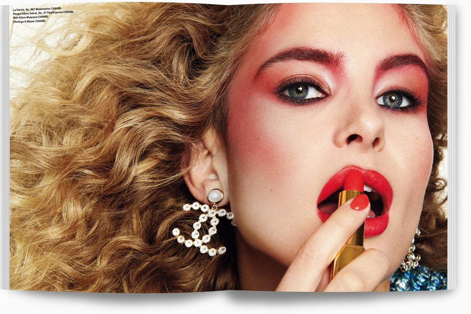 Nahaufnahme einer Frau mit lockigem blonden Haar, auffällig rotem Lippenstift, auffälligen Ohrringen, die Lippen hält sie geschlossen, während sie Lippenstift aufträgt.