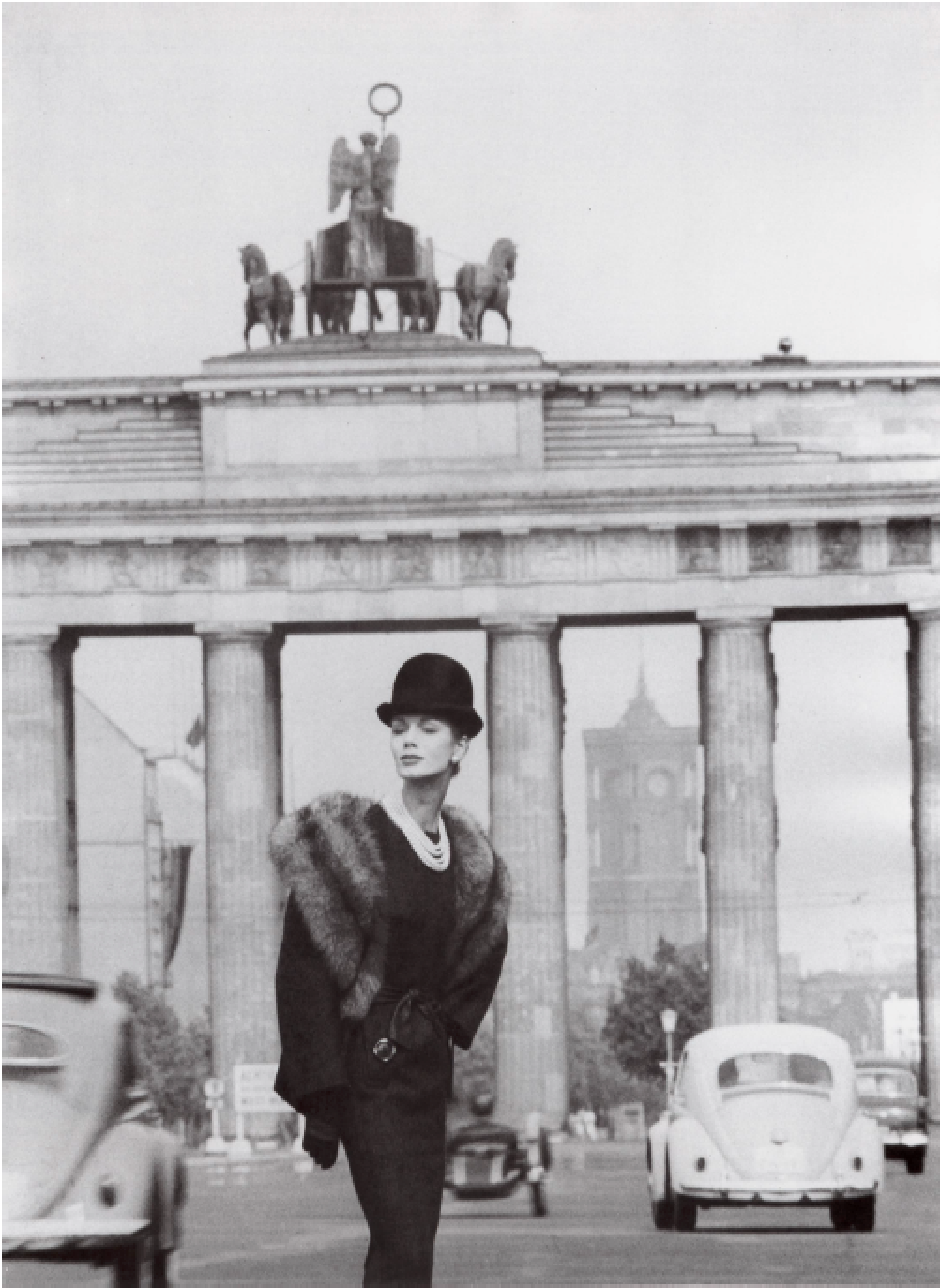 Schwarz-weiß-Foto einer Frau in eleganter Kleidung, Stehende vor dem Brandenburger Tor in Berlin, im Hintergrund Autos und der Berliner Fernsehturm.