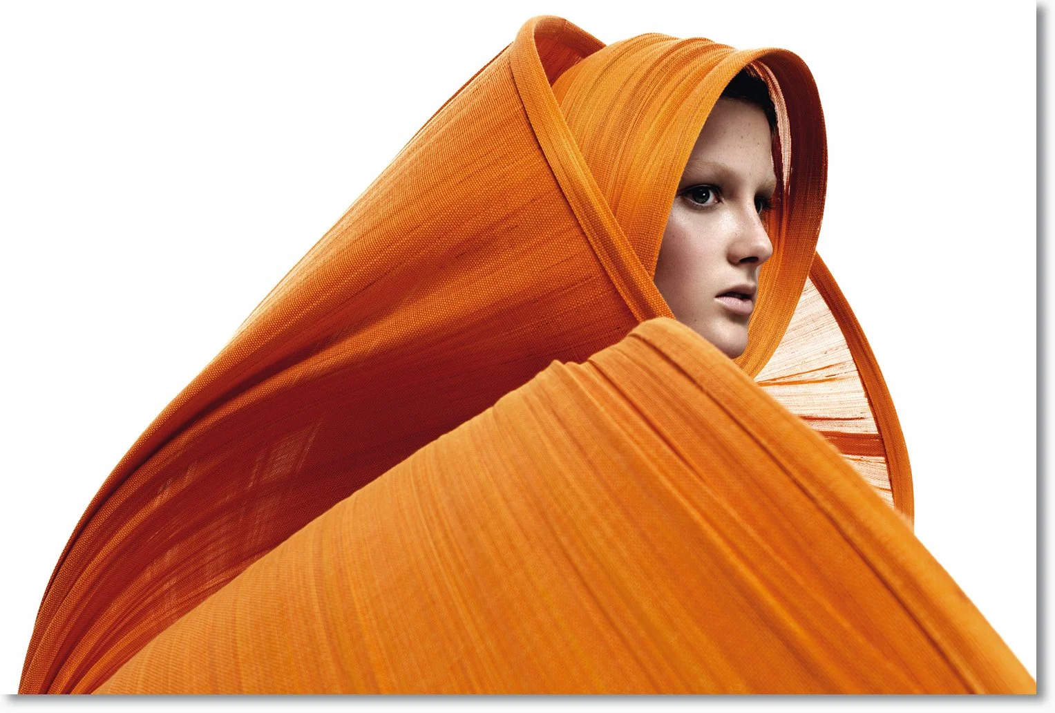 Ein junger Mensch mit hellhäutiger Haut und blonden Haaren, eingewickelt in große, orange Stoffbahnen, die an eine Blume oder ein Kunstwerk erinnern.