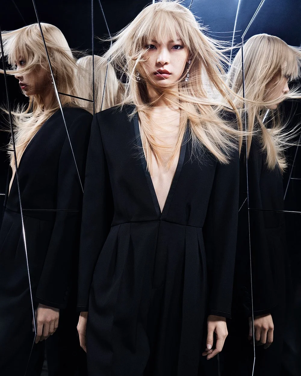 Eine Frau mit blondem Haar und dunklem Kleid steht vor einem Spiegel, der das Spiegelbild ihrer Seite zeigt, in einem Raum mit dunklem Hintergrund und Spiegelwänden.