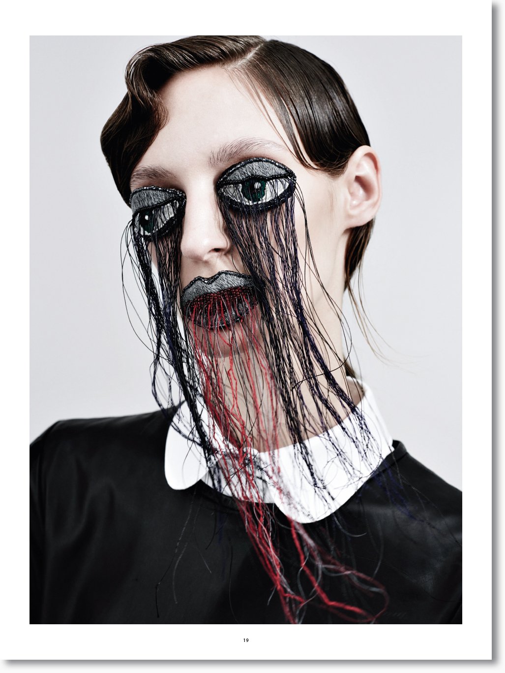 Kunstvolle Gesichtsdekoration mit aufgemalten Augen, erweiternd durch gestickte Wimpern und blutähnliche Fäden, die von den Lippen und Augen ausgehen, auf einem Model mit dunklem Haar und schwarzem Outfit.