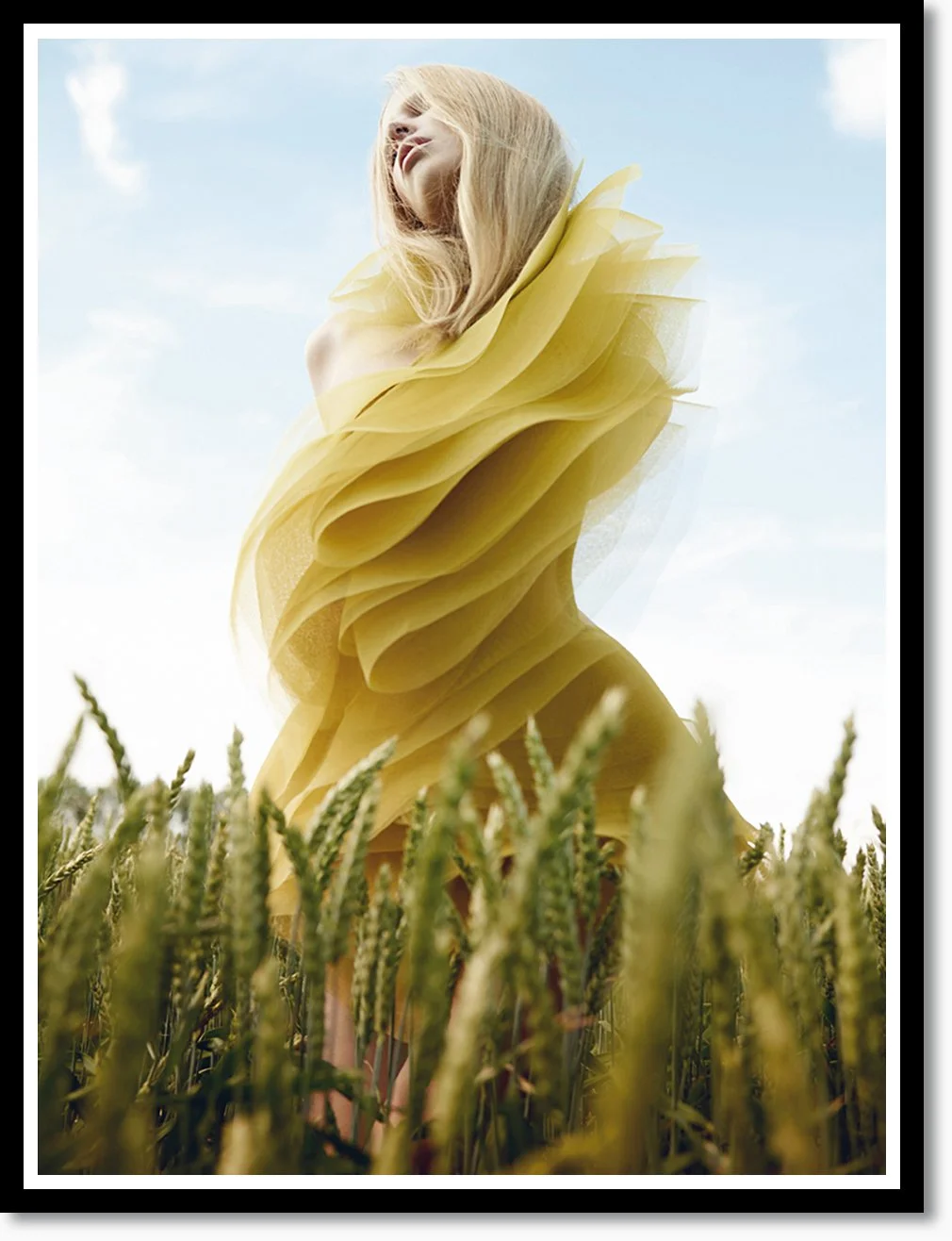 Eine Frau in einem gelben, zarten Kleid steht in einem Feld aus Weizen. Der Himmel ist blau mit wenigen Wolken.