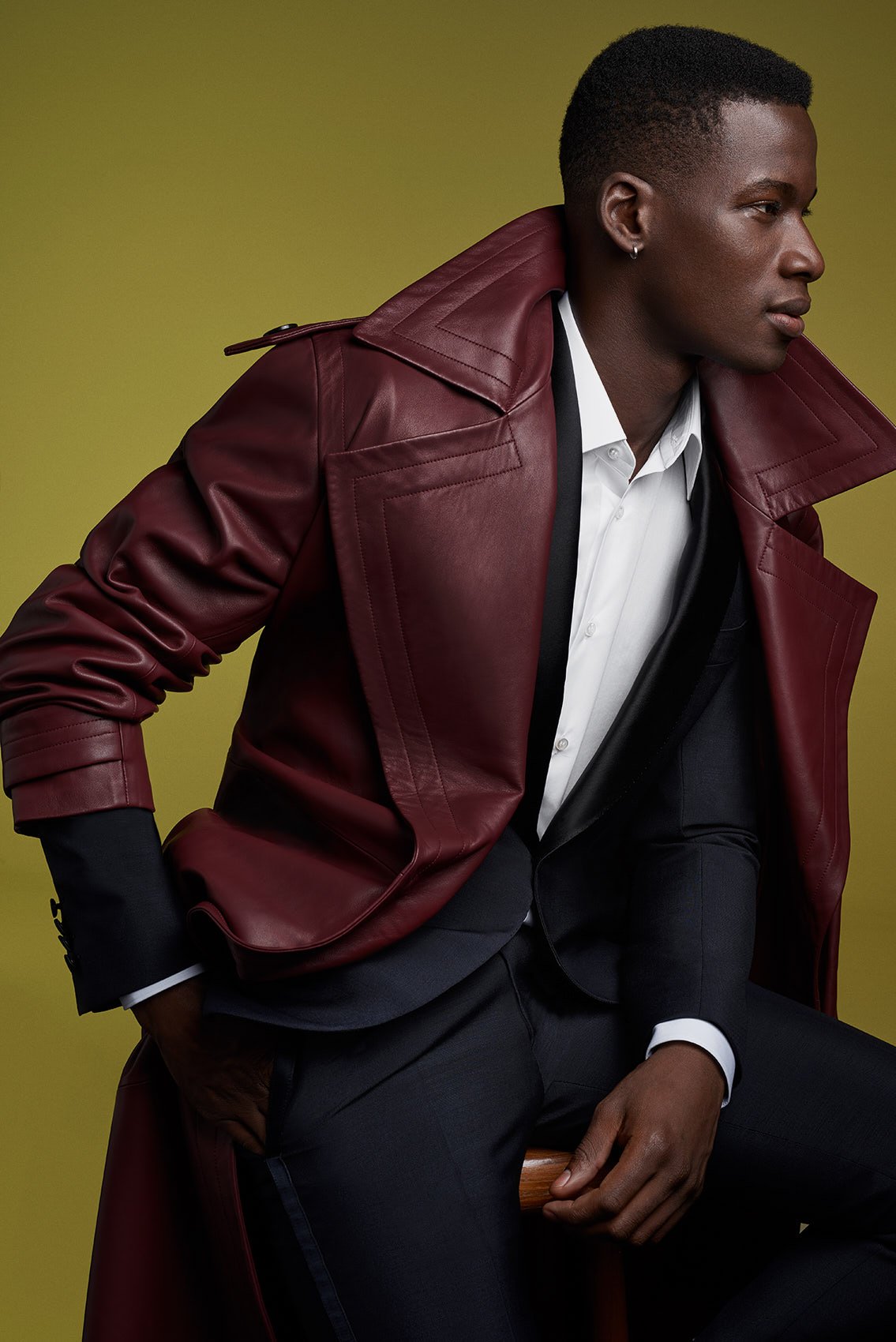 Junge schwarze Person trägt eine weiße Hemd, schwarzen Anzug, rote Lederjacke und sitzt auf einem Holzstuhl vor einem gelben Hintergrund.