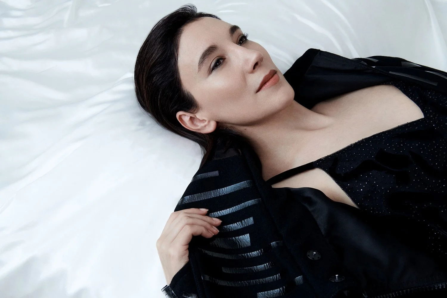A woman with dark hair lying on a white bed, wearing a black top and a black jacket.